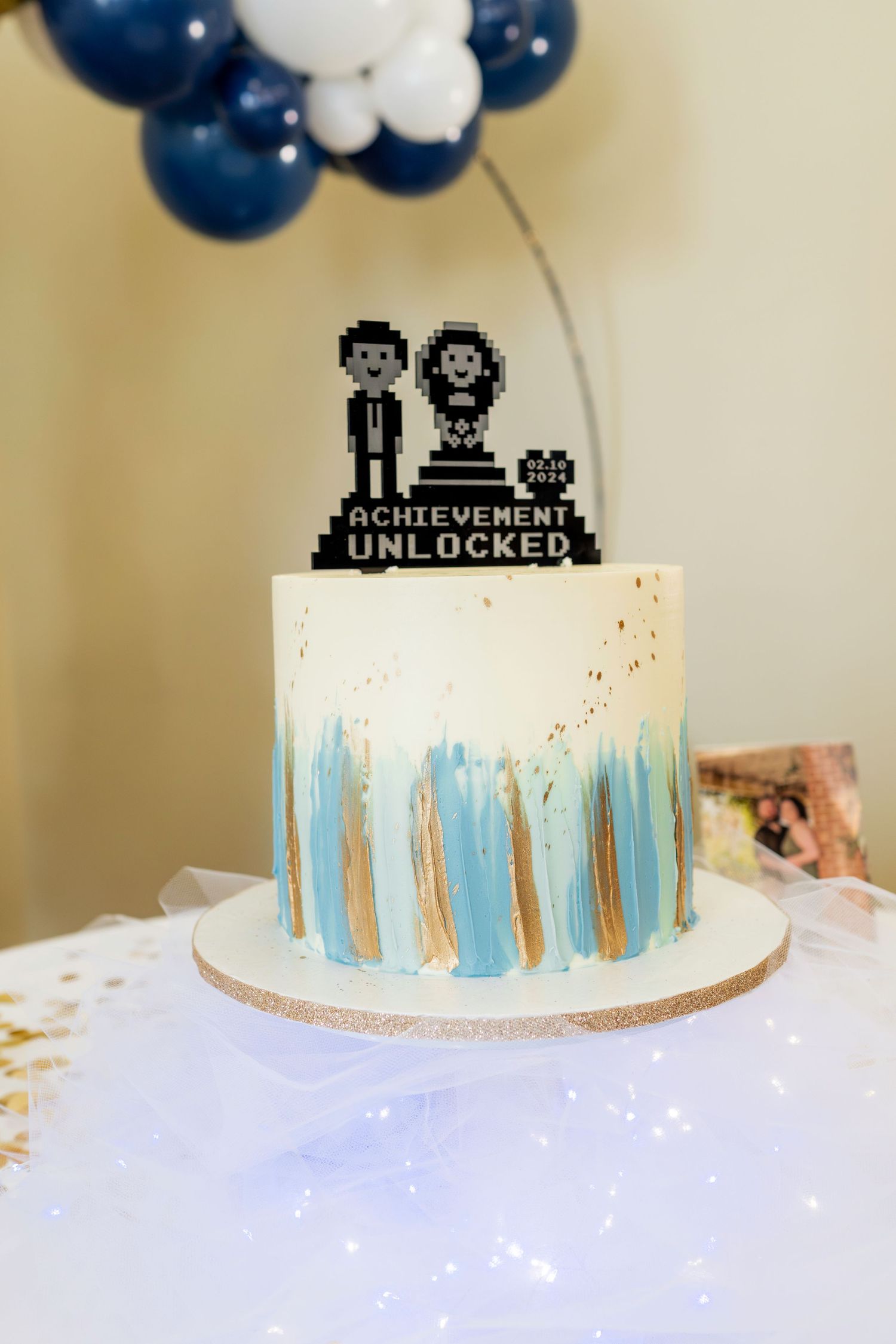 Gaming themed wedding cake with pixel art topper and blue watercolor design against balloon backdrop.