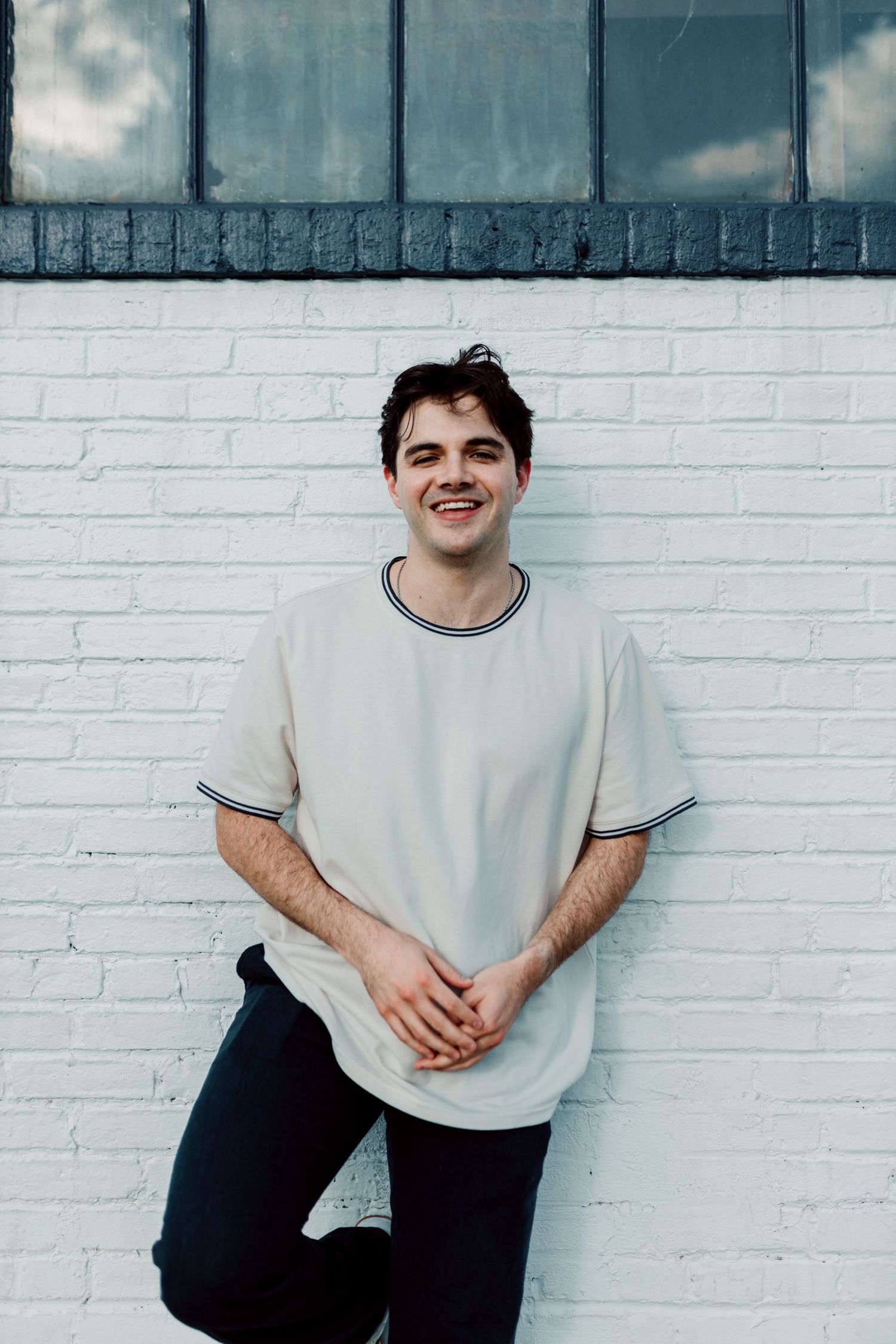 A casual portrait against a white brick wall with industrial windows above, wearing a light colored t-shirt with navy trim.
