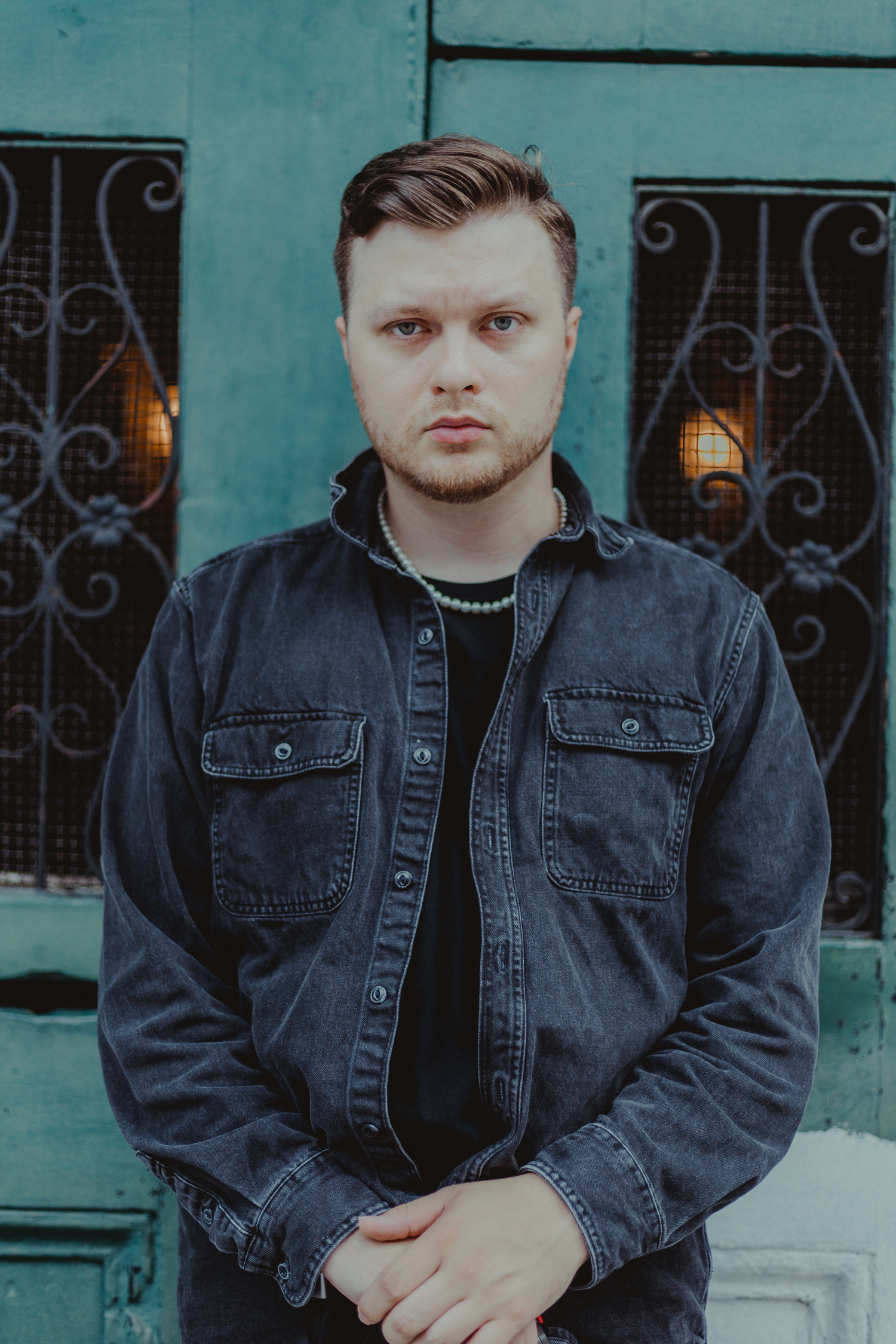 Portrait of someone in a dark denim jacket against a turquoise wall with decorative ironwork.