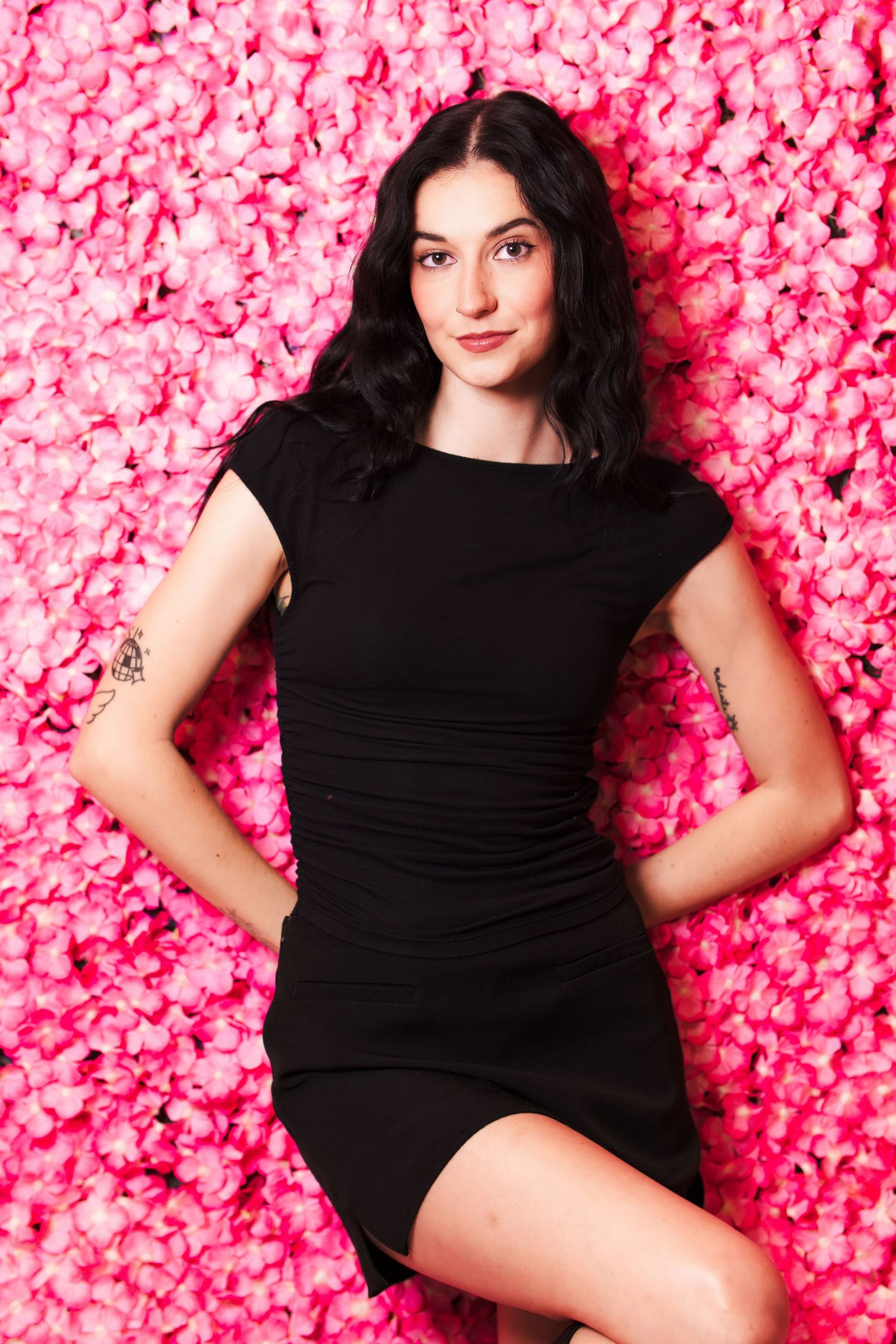A woman in a little black dress poses against a vibrant pink floral backdrop.