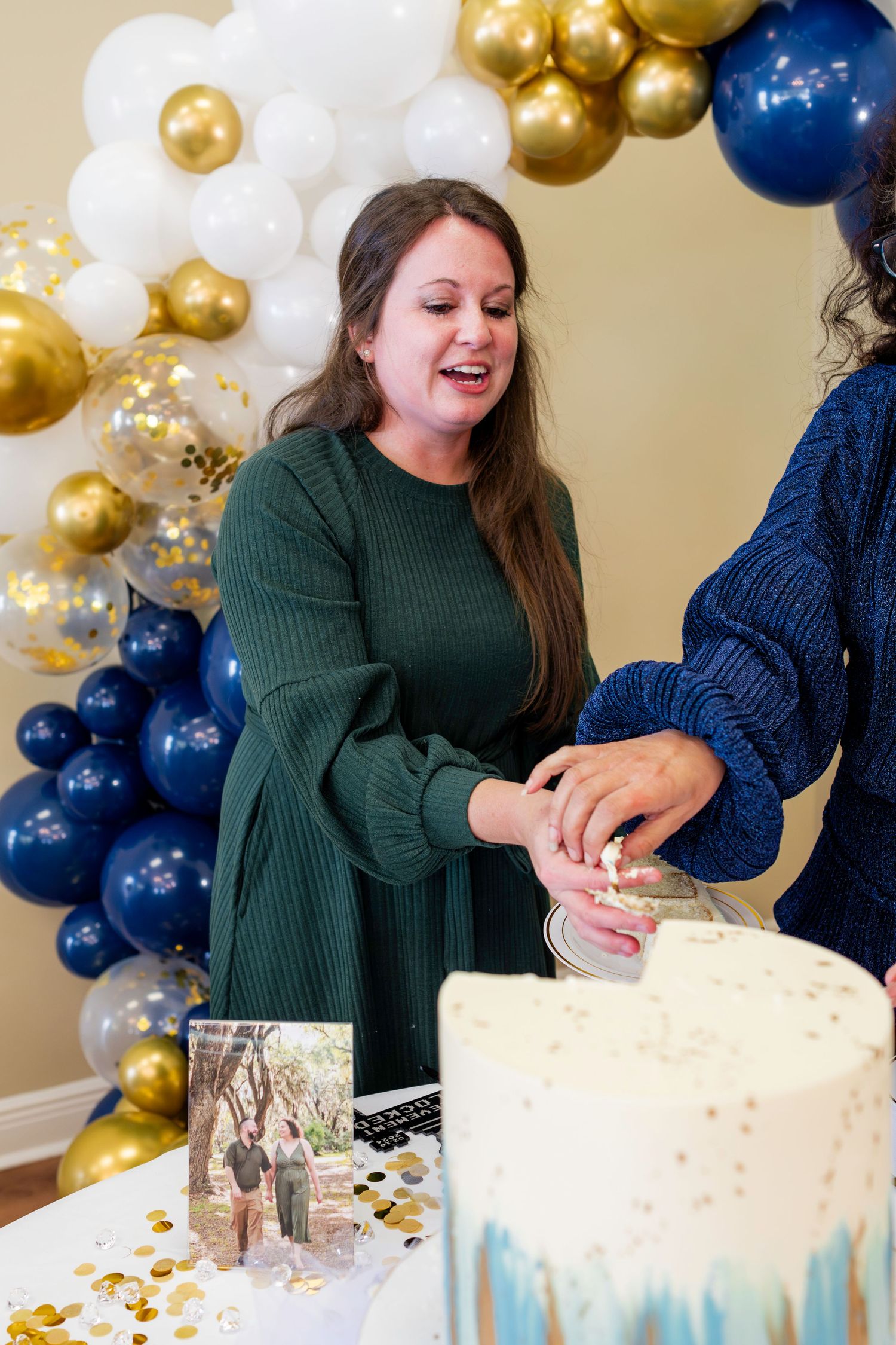 A celebration party decorated with gold and navy blue balloon arch and a cake being cut.