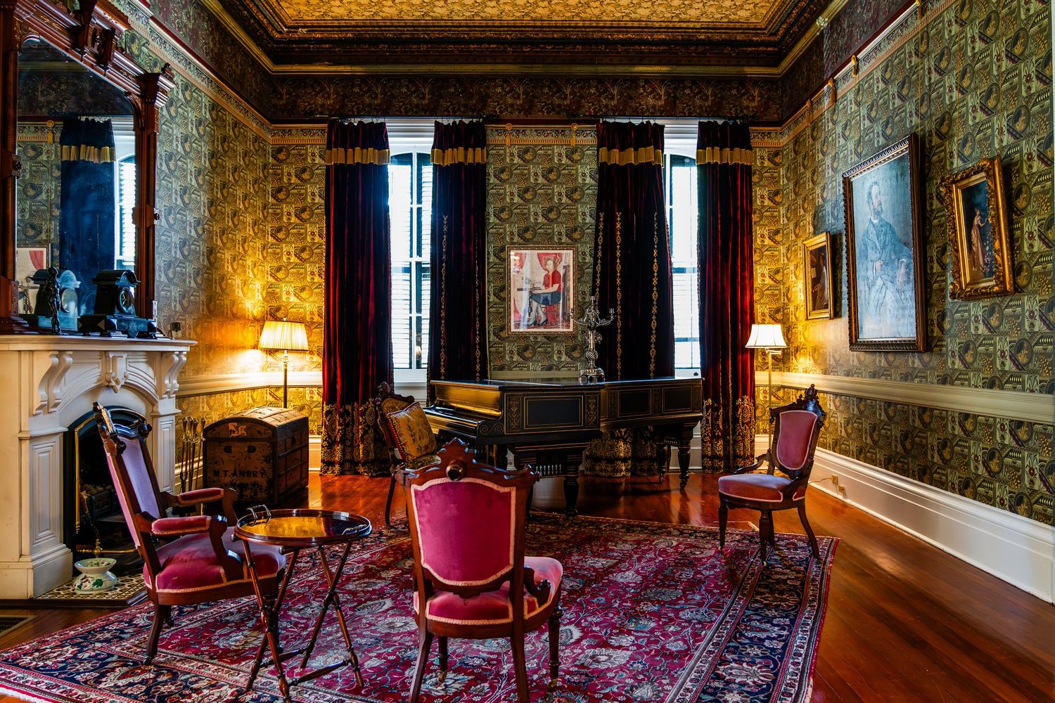 Victorian parlor room with ornate wallpaper, red velvet chairs, oriental rug, and warm lamp lighting creates elegant ambiance.