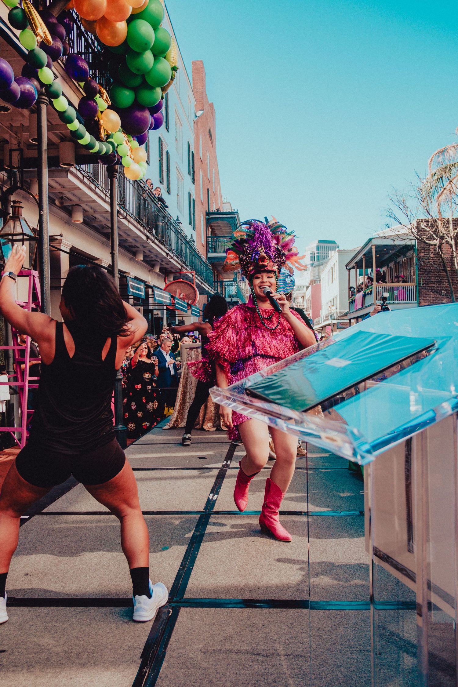 Street performer in pink feathers dances during Mardi Gras celebration under balloon decorations.