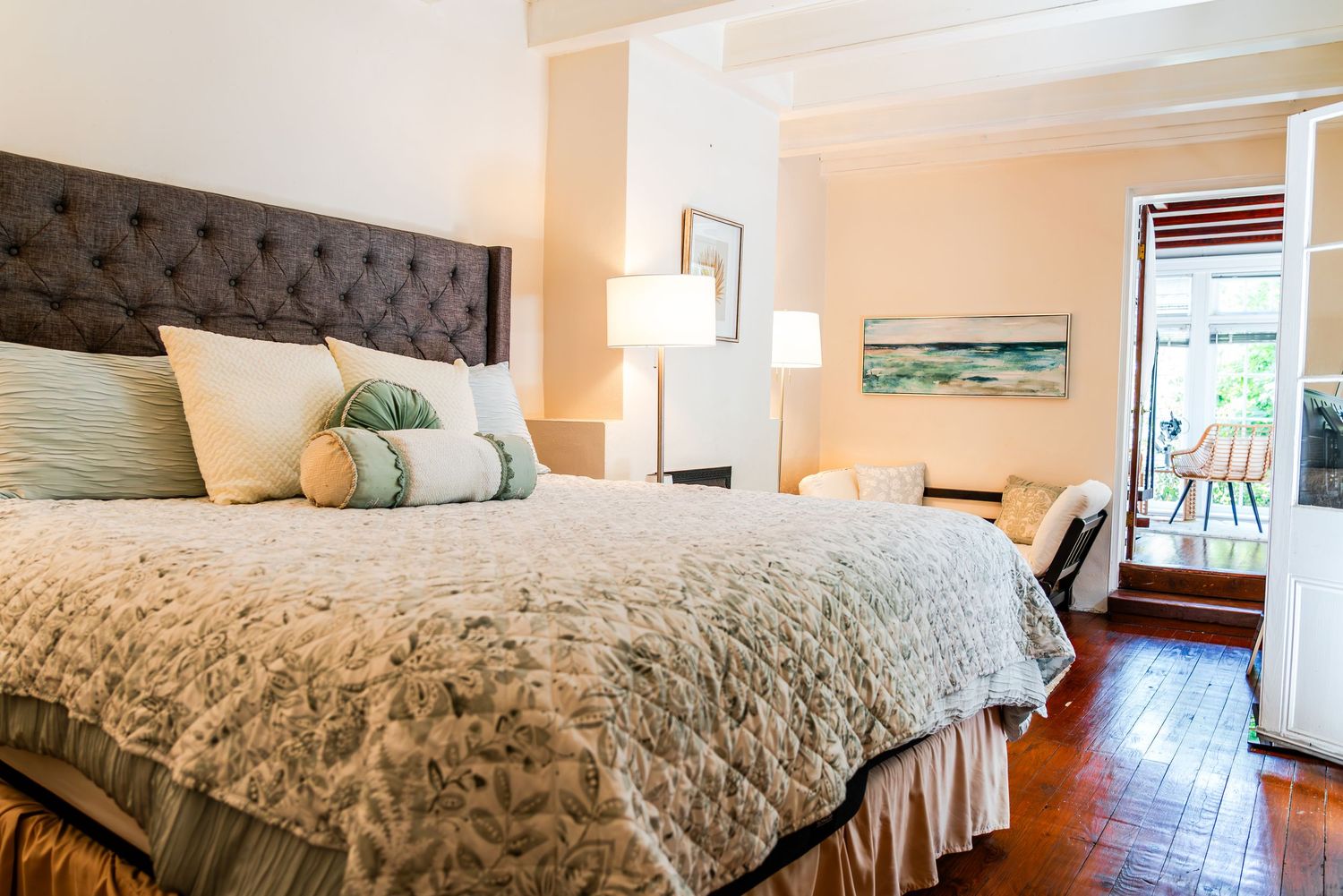 A cozy bedroom with a gray tufted headboard, plush bedding, and wooden floors leading to a private balcony.