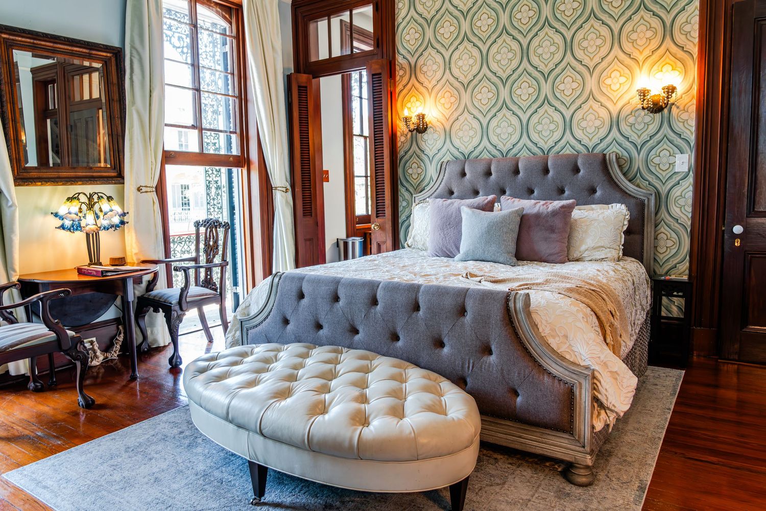 Luxurious bedroom featuring a tufted gray velvet bed, curved chaise lounge, and elegant patterned wallpaper with hardwood floors.