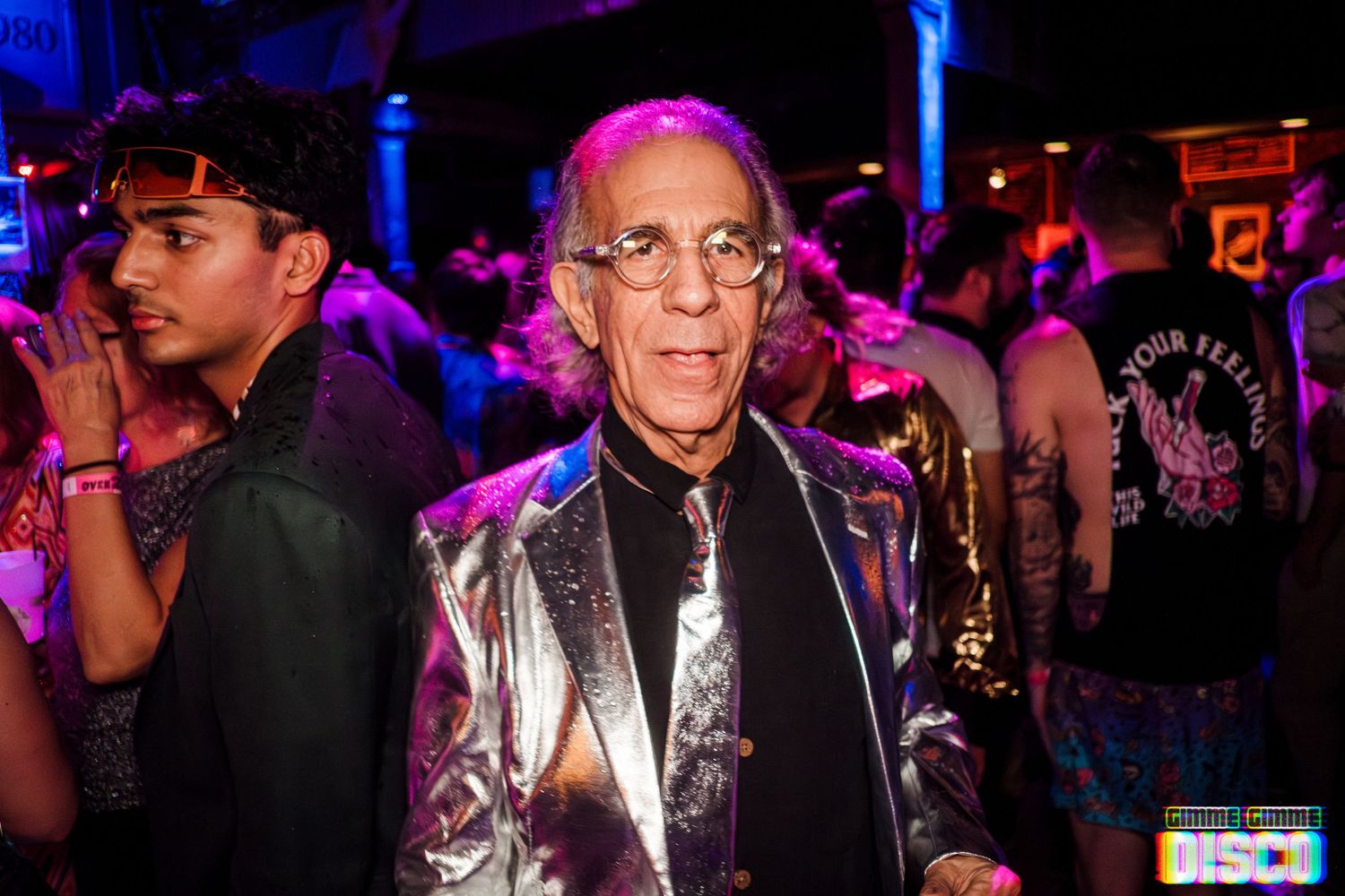 A party-goer in a metallic jacket stands among the crowd at a nightclub event.