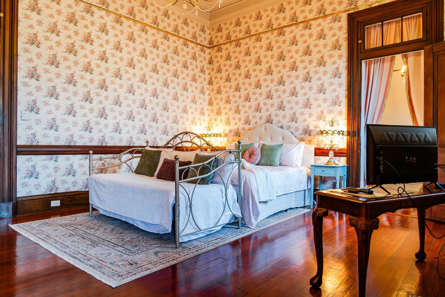 Elegant Victorian bedroom with ornate wallpaper, antique iron bed frame, and crystal chandelier in muted tones.