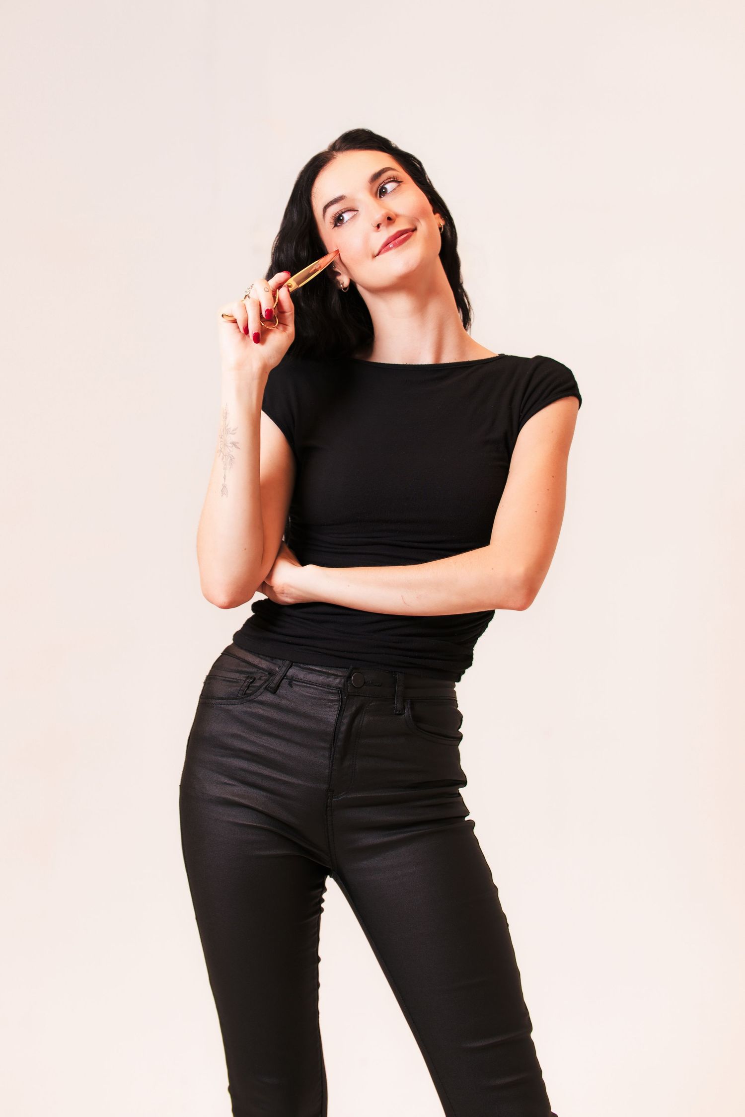 Someone in an all-black outfit stands thoughtfully against a light background while looking up and smiling.