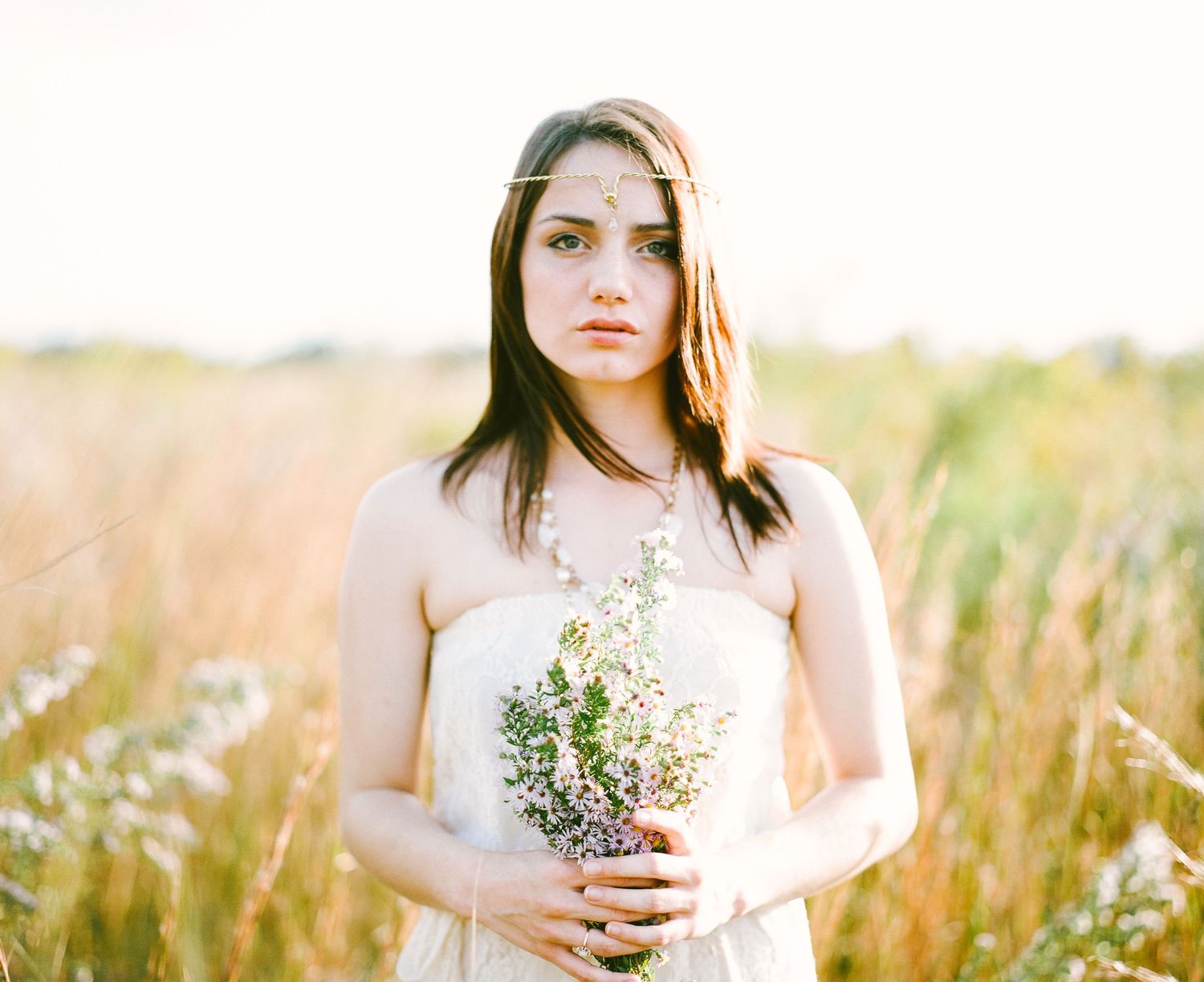 A person in a white dress stands in a golden wheat field holding a delicate bouquet of wildflowers at sunset.