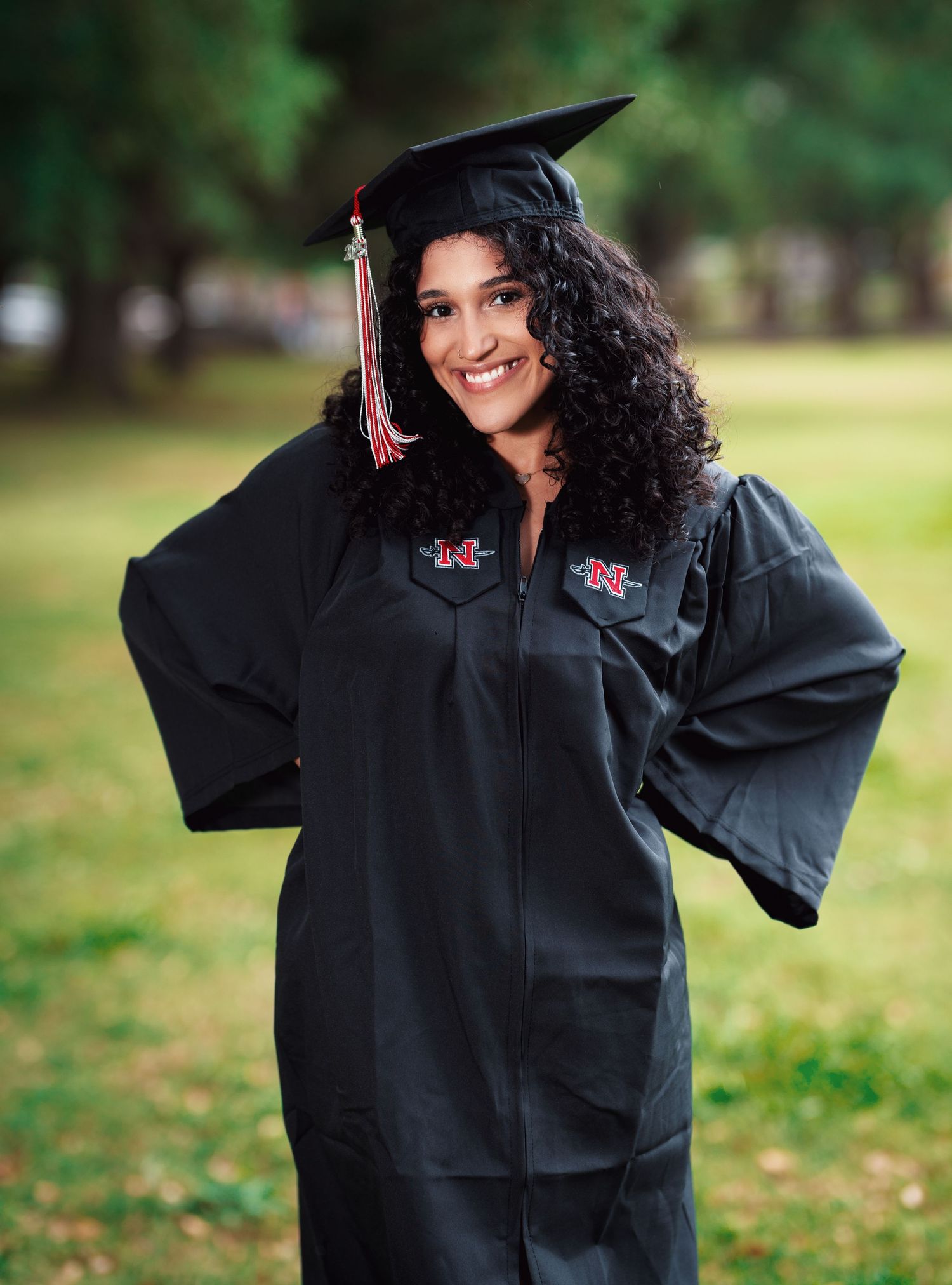 Graduate in black cap and gown with red tassel smiling outdoors.