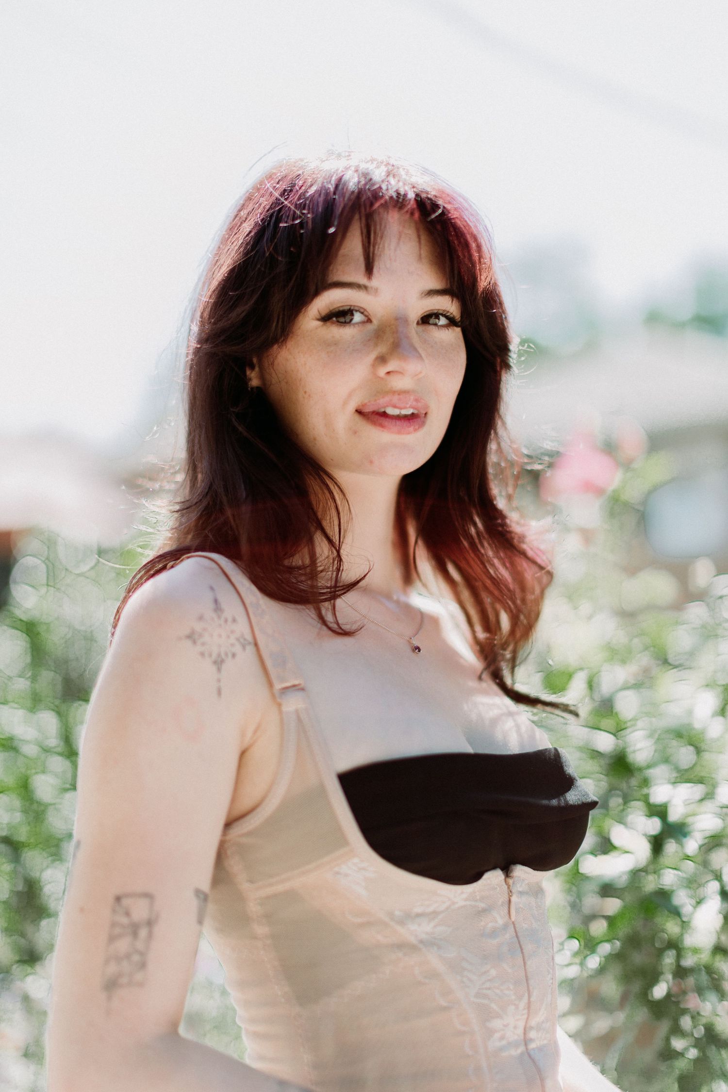 Portrait of someone in a light dress with dark trim outdoors, with sunlit greenery and bokeh lighting in the background.