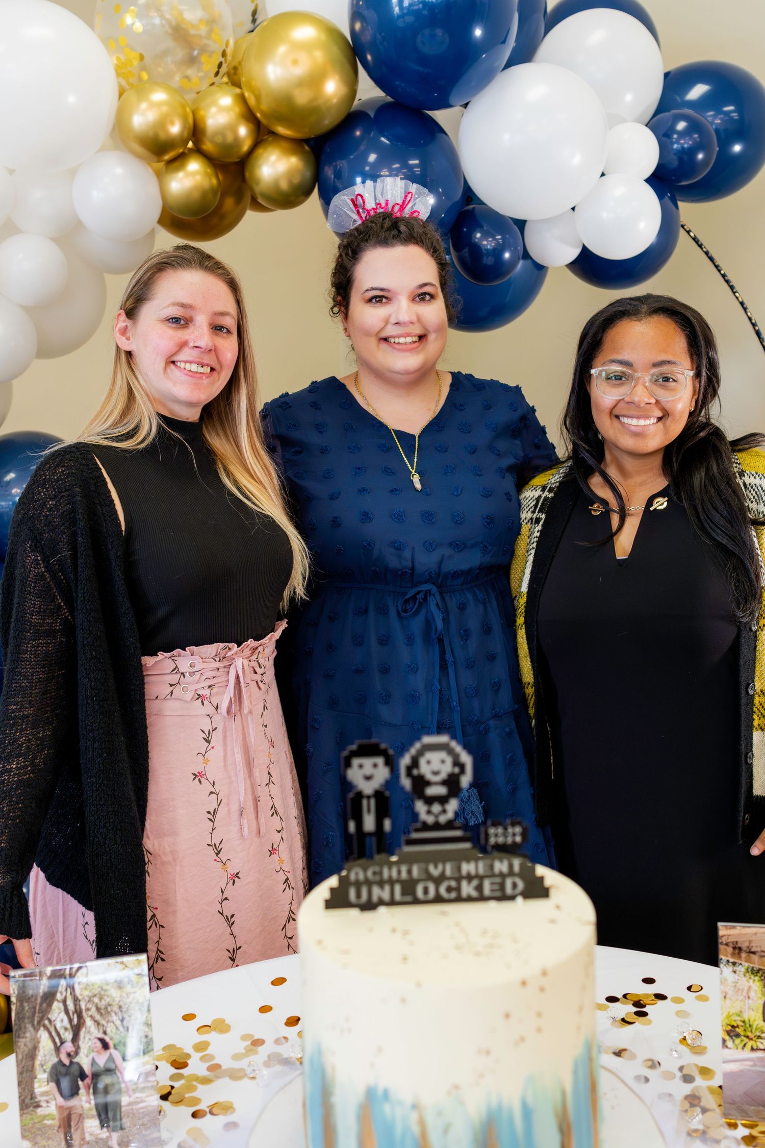 A celebration with balloons features three people posing by a cake with Achievement Unlocked decorations.