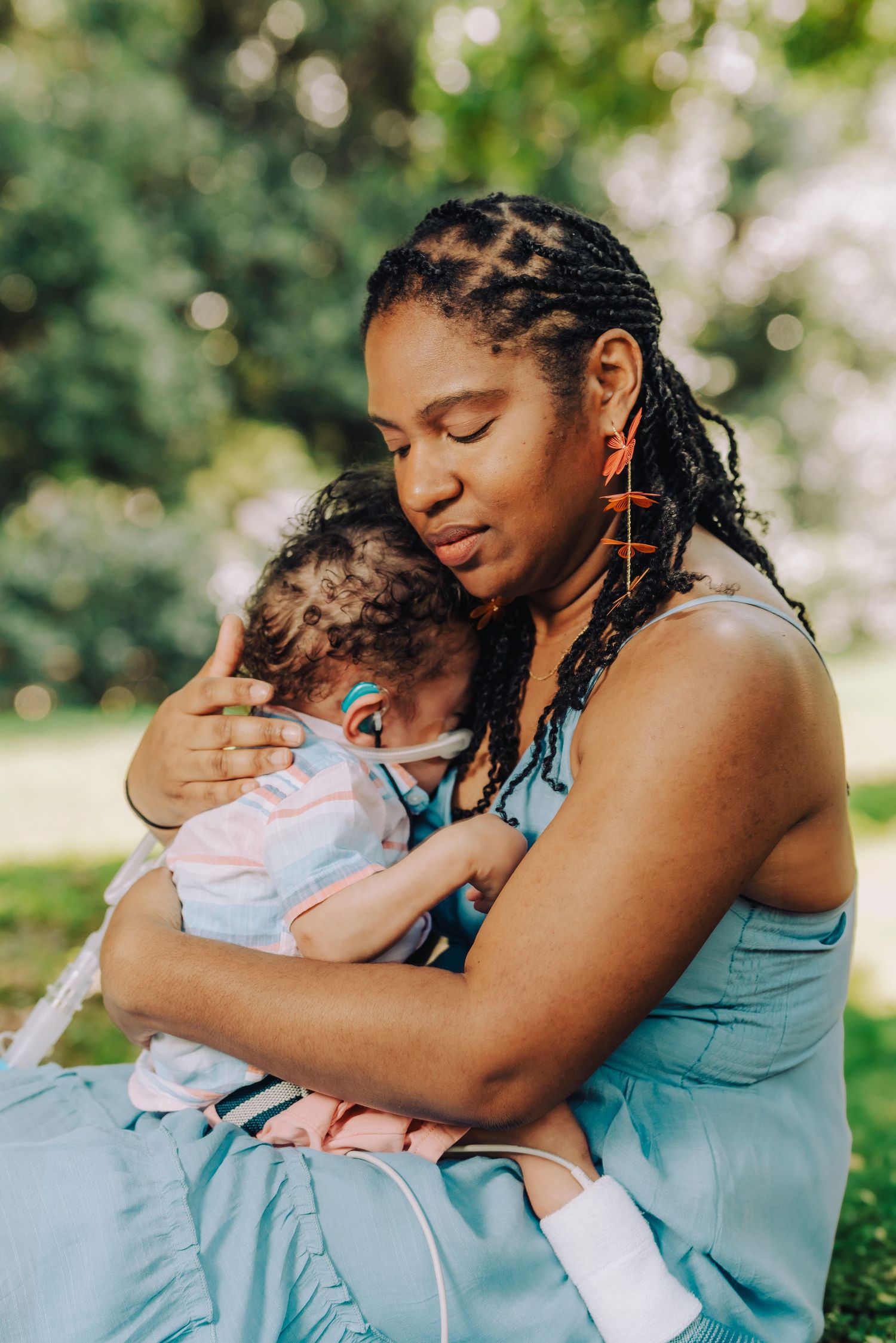 Parent in light blue dress embraces infant outdoors in sunlit setting with soft bokeh background.