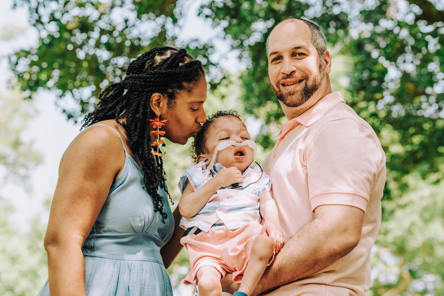 Three family members share joy and laughter outdoors on a sunny day surrounded by green trees.