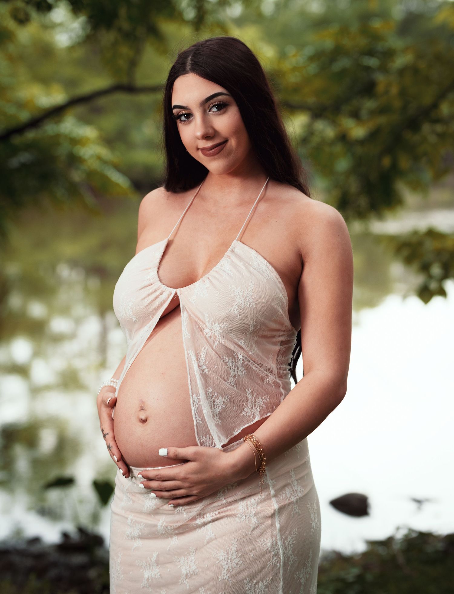 A maternity photoshoot in nature featuring a white lace dress and ethereal lighting by a lake.