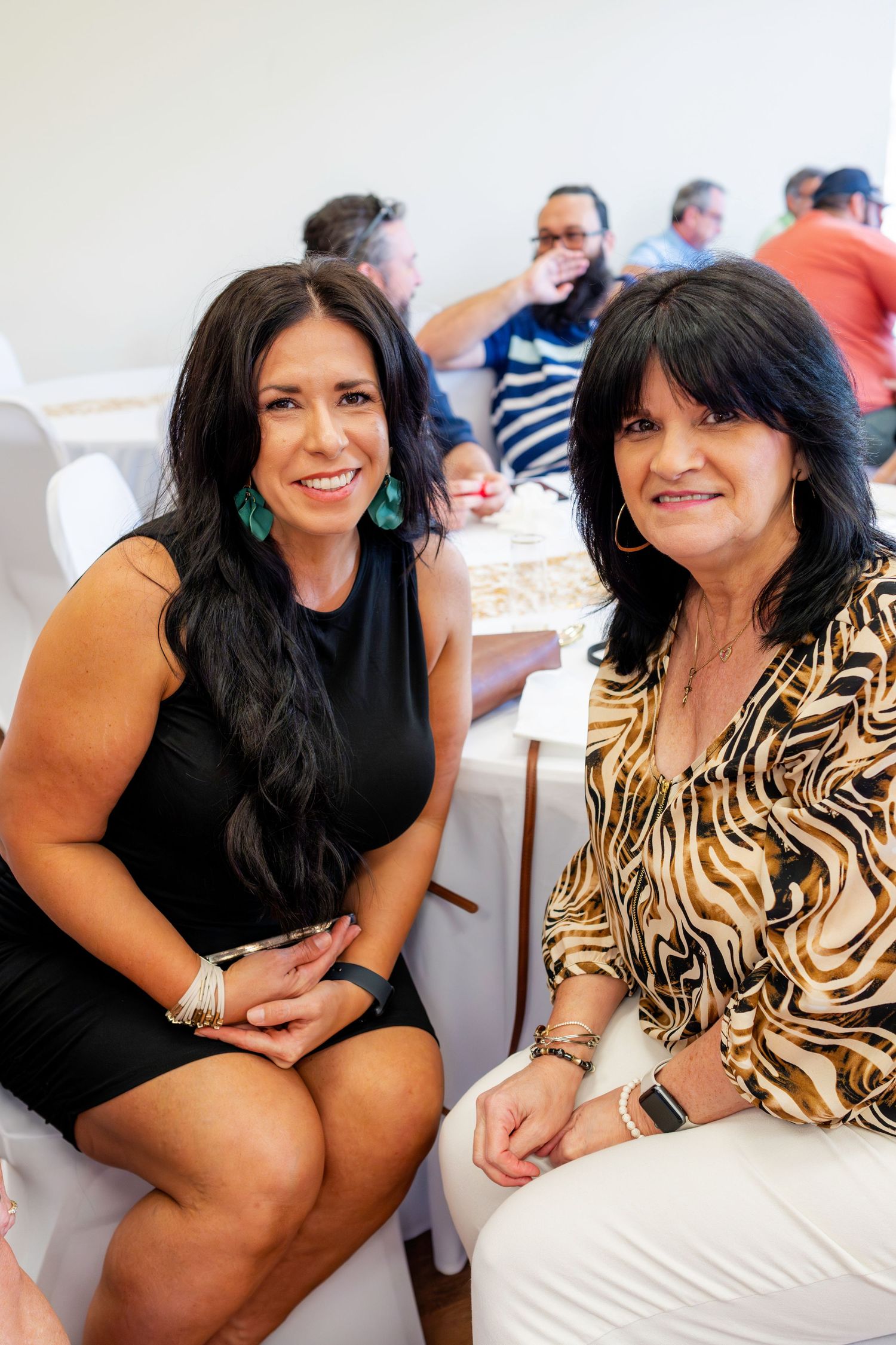 Two women in stylish attire smile while seated at a festive party celebration.