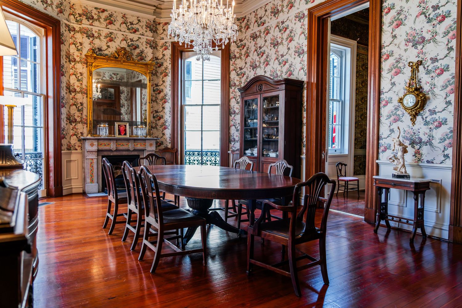 Elegant Victorian dining room with floral wallpaper, crystal chandelier, dark wood furniture and polished hardwood floors.