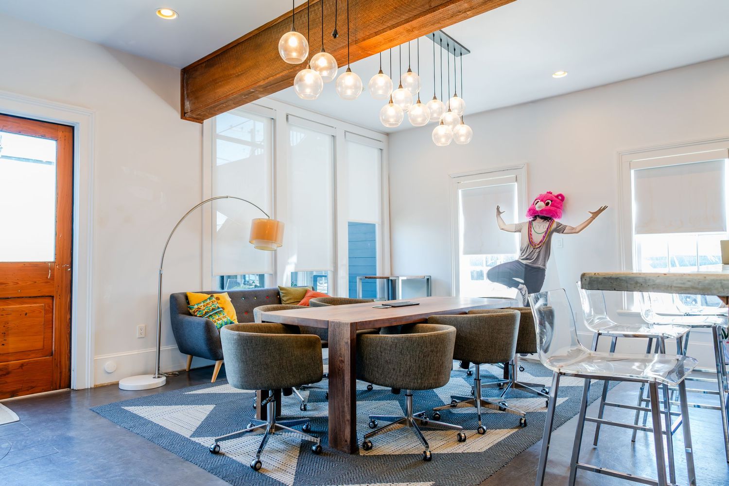 Modern dining room features wood table with teal chairs, hanging glass globe lights and exposed wooden ceiling beam.