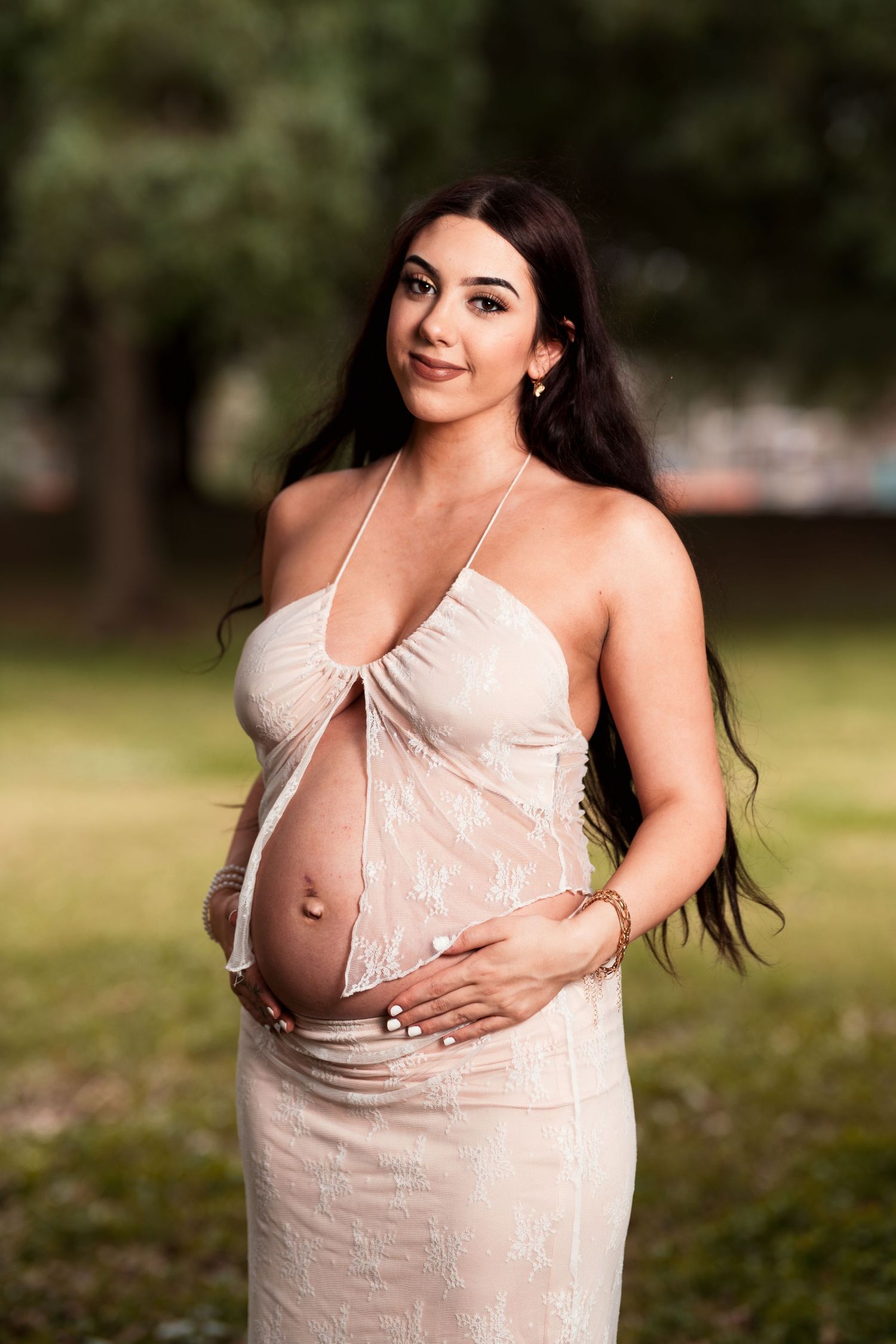 Outdoor maternity portrait showing a pregnant woman in a cream-colored dress smiling at the camera.