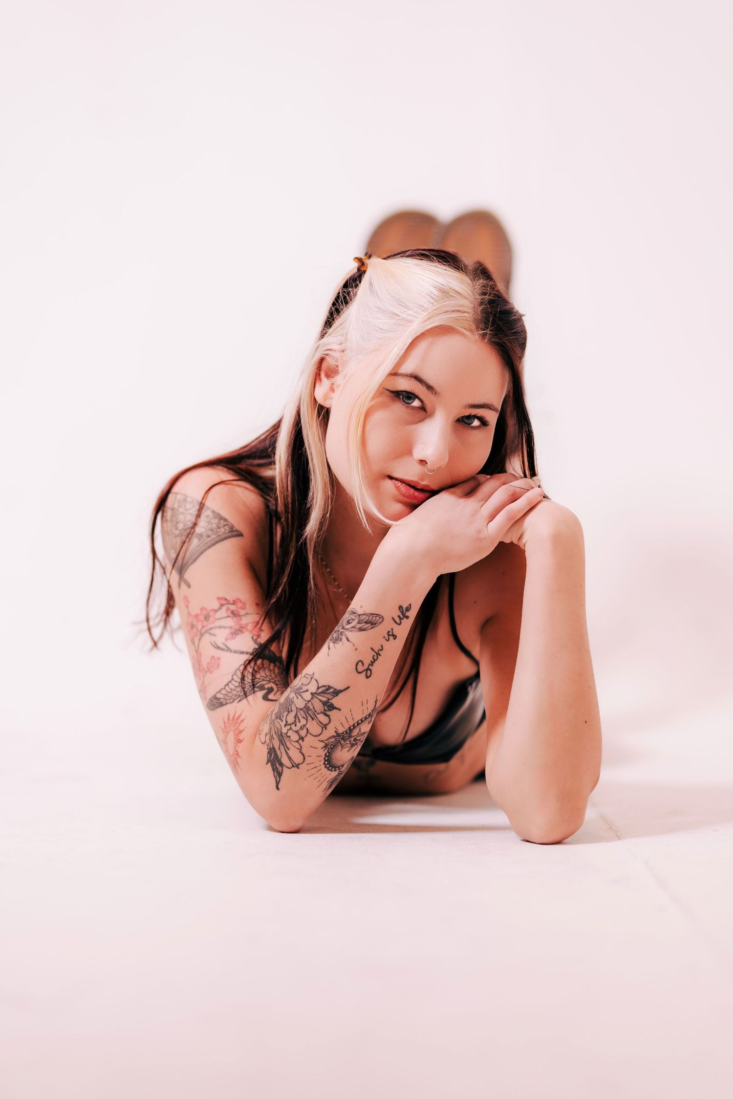Person with arm tattoos laying on a white surface in a boudoir style pose wearing a black top against pink backdrop.