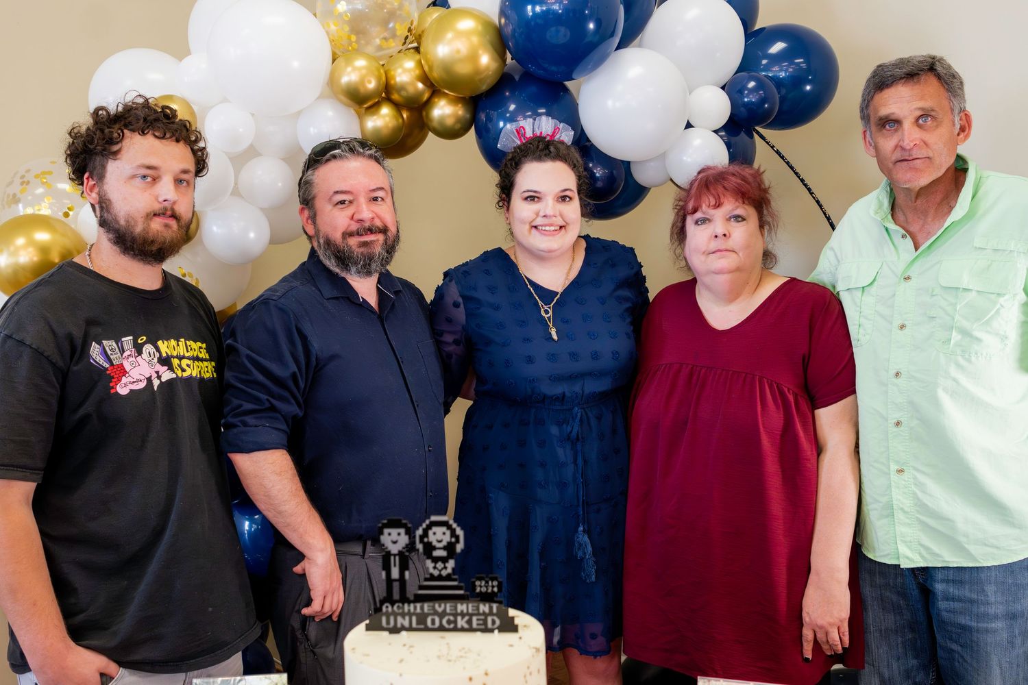 A group photo at a celebration with a cake and decorative blue, white and gold balloons in the background.