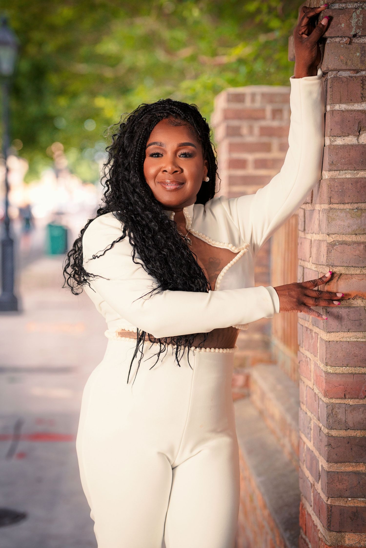 Woman in a white jumpsuit with long curly hair poses against a brick wall on a city street.
