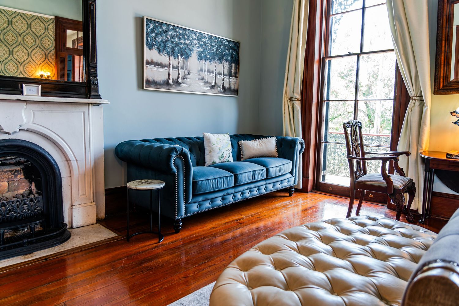 A luxurious living room features a teal velvet chesterfield sofa, ornate fireplace, and polished hardwood floors.