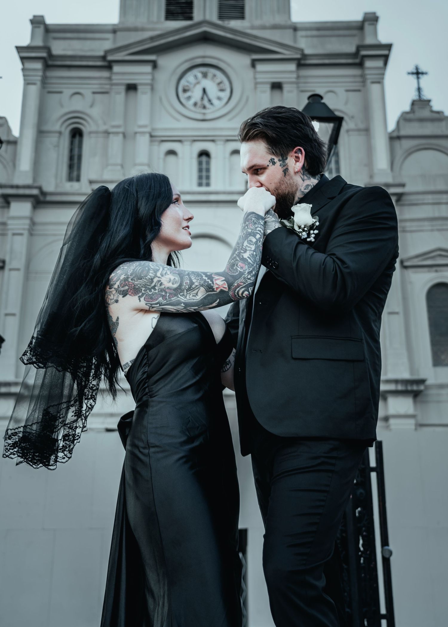 A couple in dark formal attire share an intimate dance pose outside a historic building with cathedral architecture.