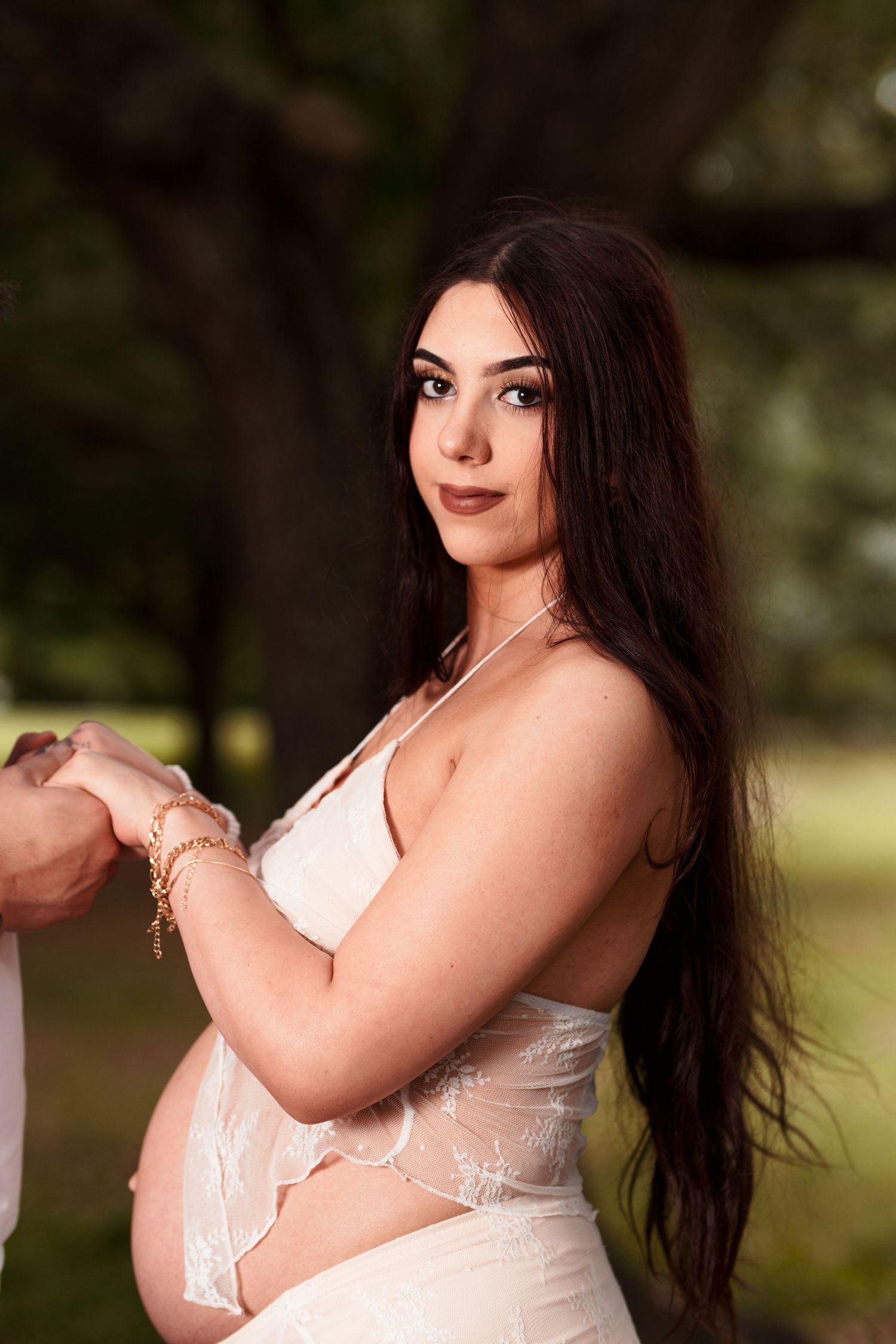 An outdoor maternity portrait with long dark hair and white lace dress against a natural background.
