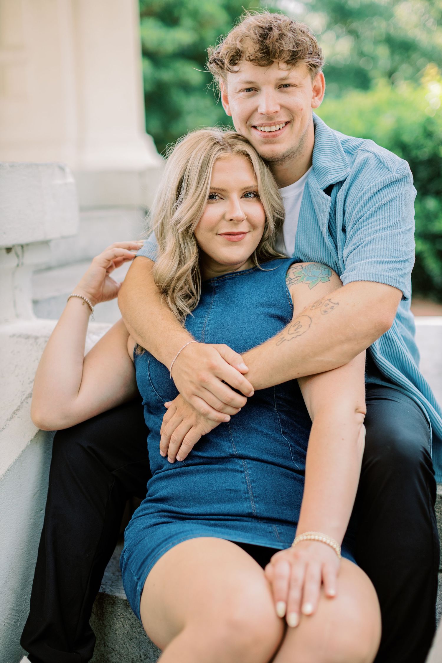 A couple embraces outdoors while sitting on stone steps, with her in a blue dress and him in casual attire.