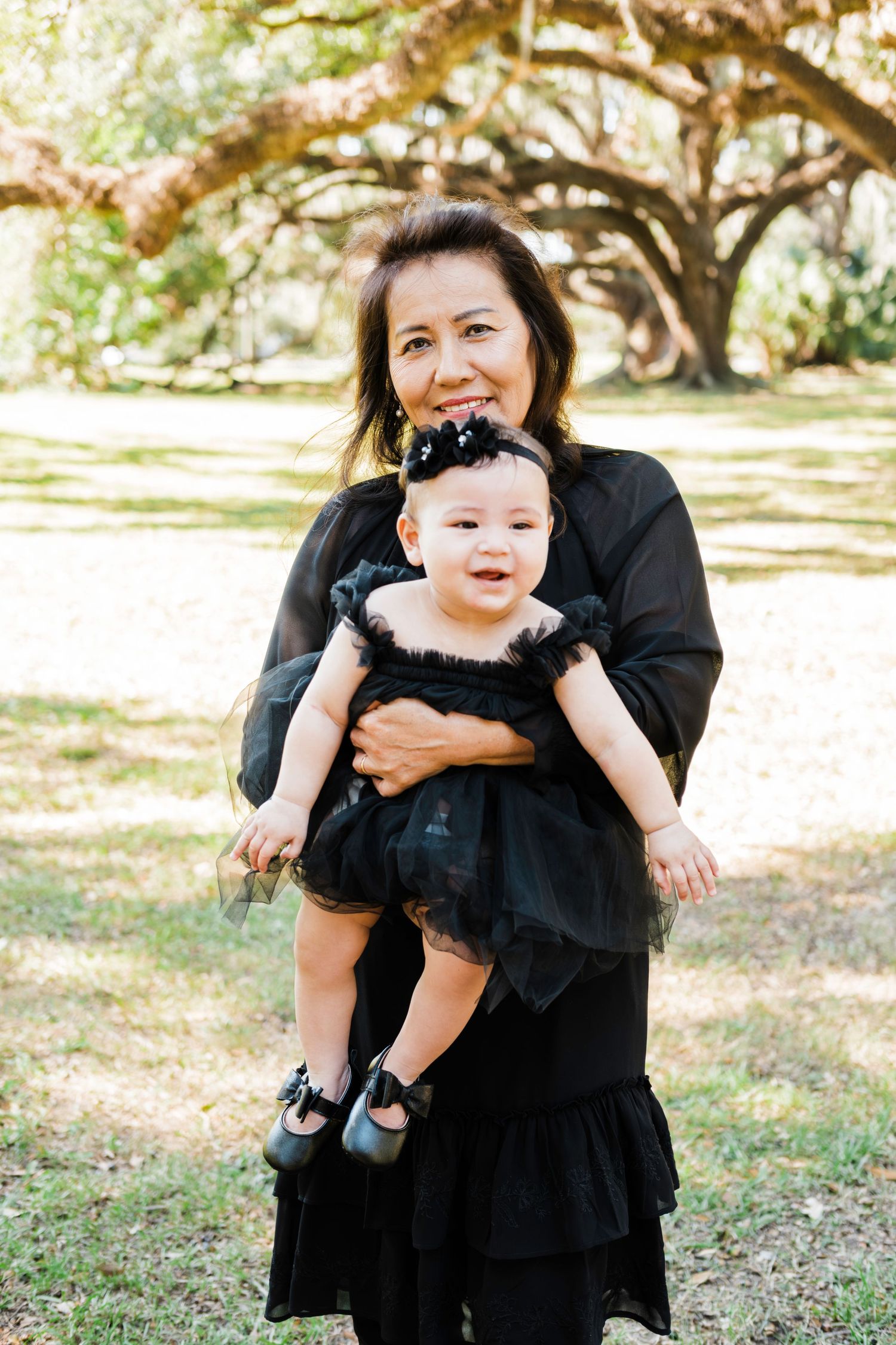 A heartwarming moment captured between parent and baby in black formal attire under oak trees.