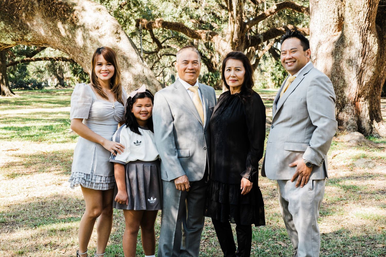 A family poses together outdoors in a park setting with trees in the background on a sunny day.