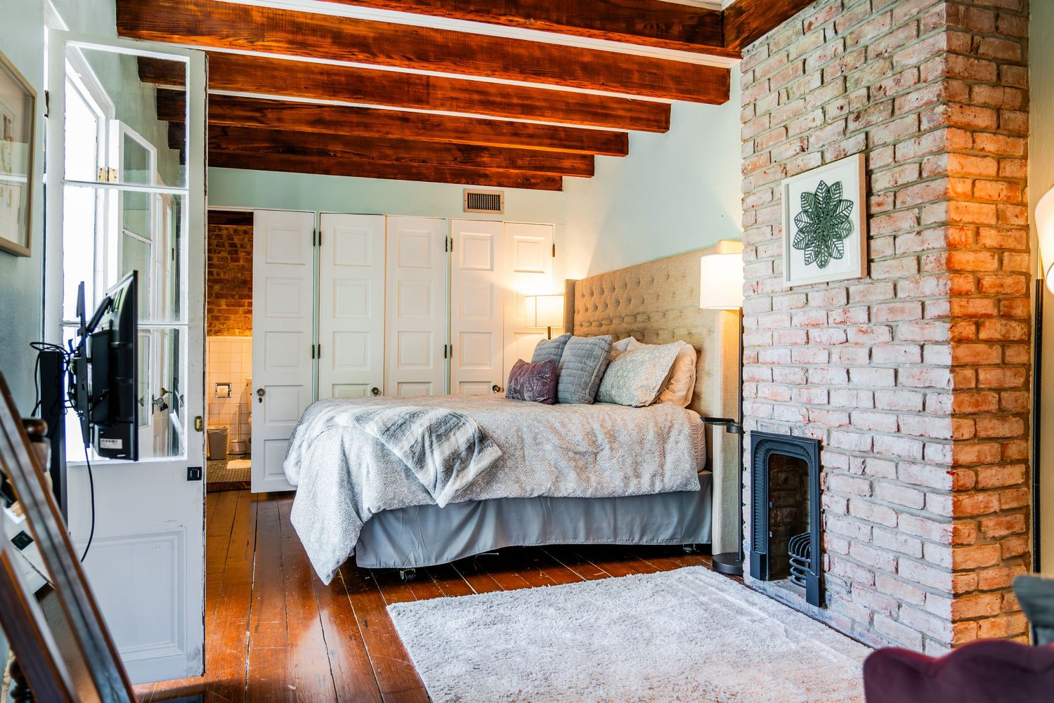 Modern bedroom features exposed brick wall, wooden beams, plush upholstered bed, and natural light from white french doors.