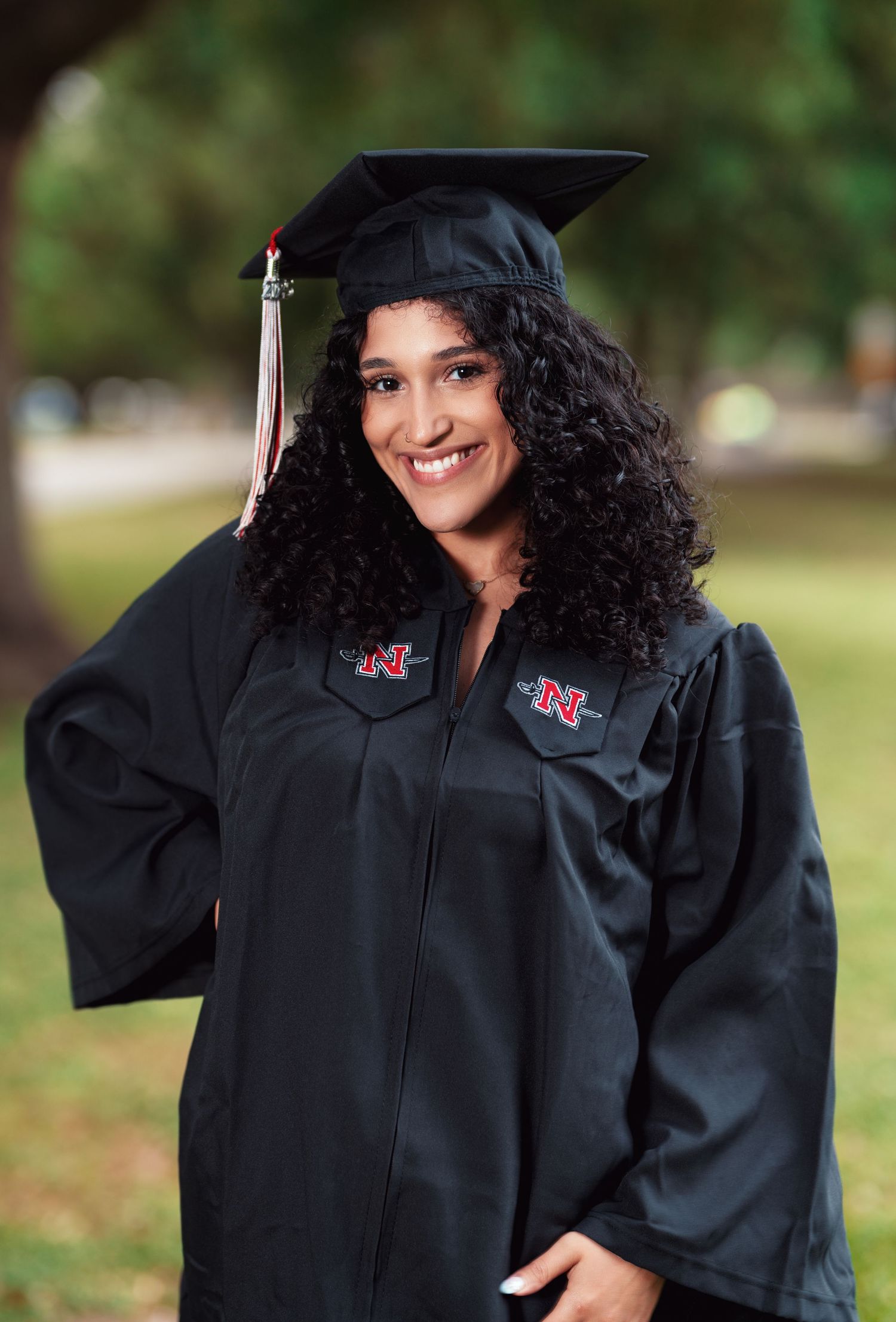 Graduate wearing academic regalia smiles brightly against blurred outdoor background.