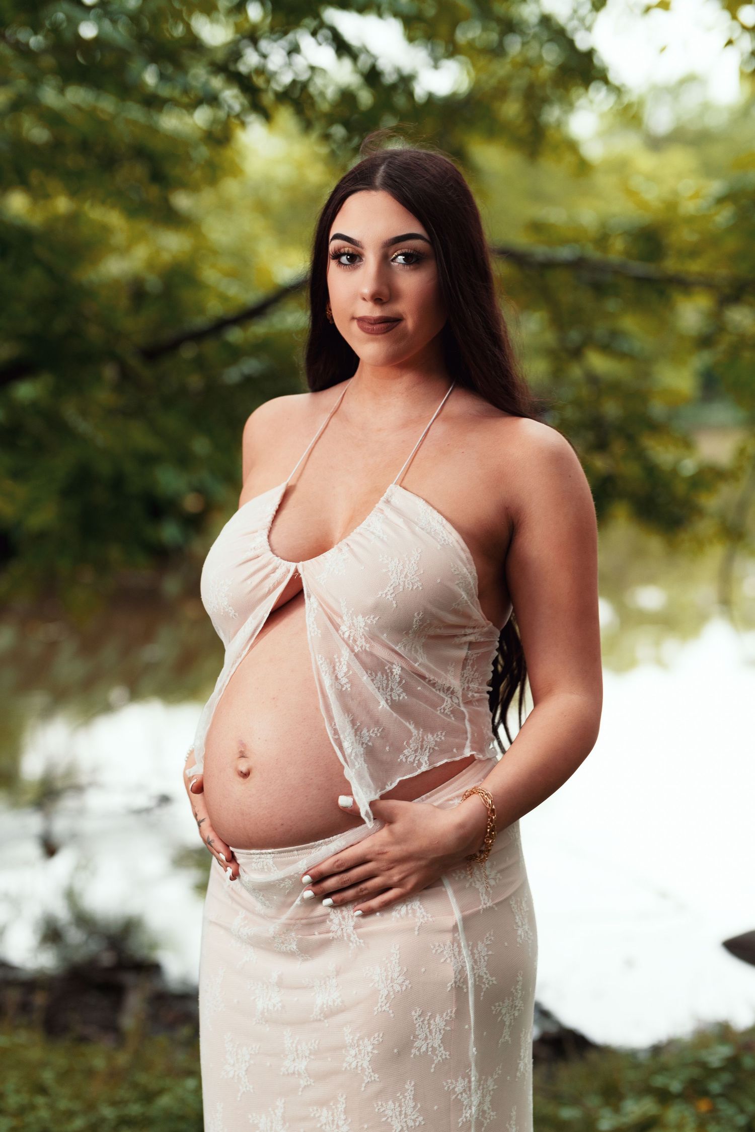 Maternity portrait of an expectant mother in a white lace dress standing outdoors near water in natural lighting.