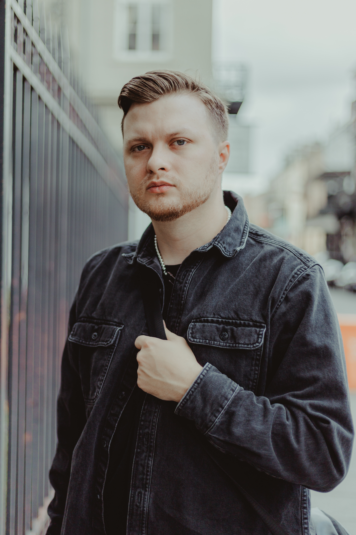 Portrait of someone in black denim jacket against urban background with metal fencing.