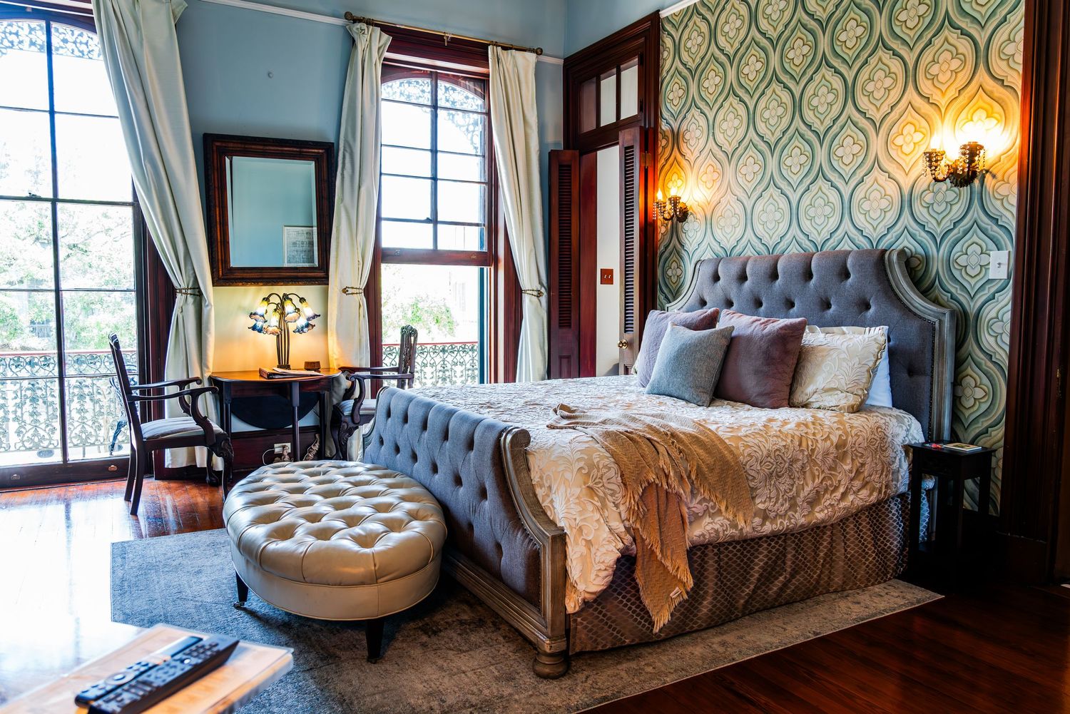 Elegant bedroom with tufted blue headboard, ornate wallpaper, floor-to-ceiling windows, and vintage-style decor.