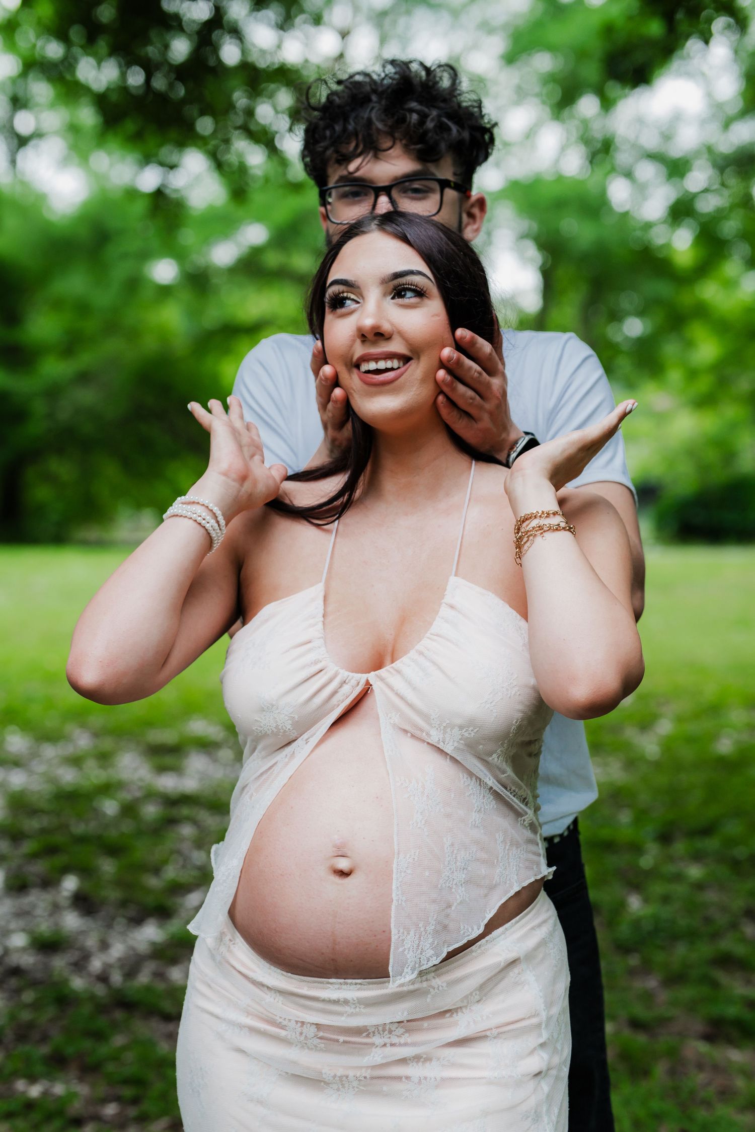 Candid maternity moment captured of couple sharing joy in green outdoor setting.