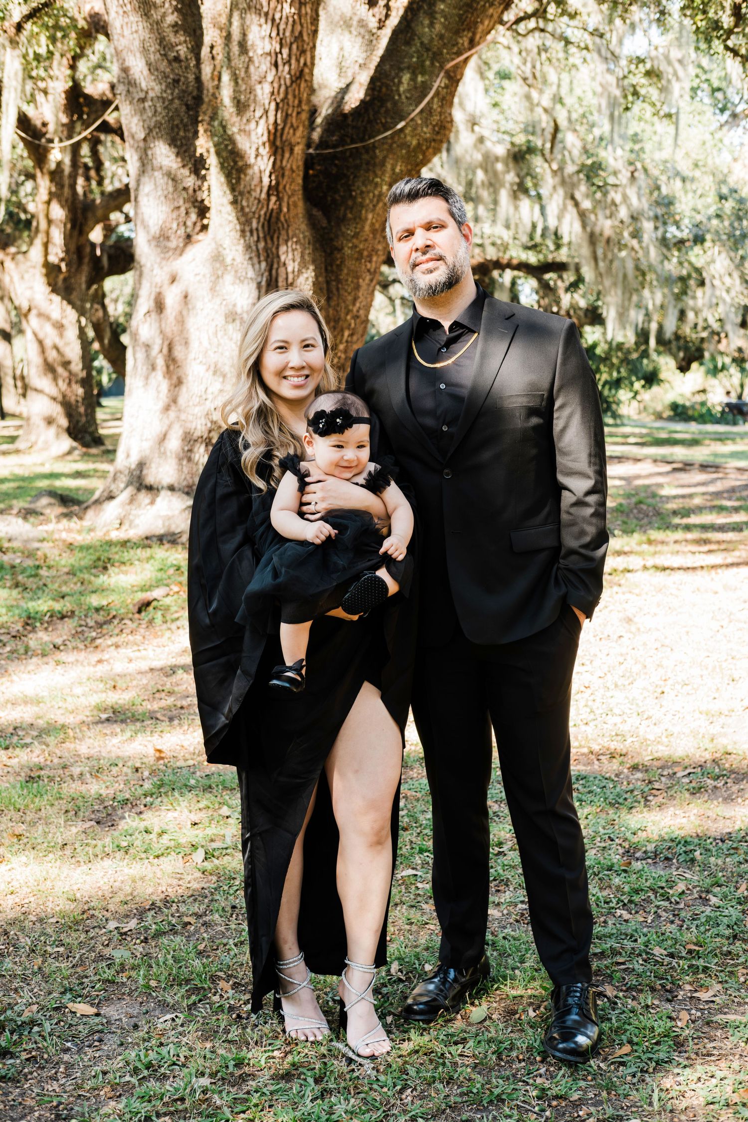 A couple in black formal wear holds their baby while standing under Spanish moss trees.