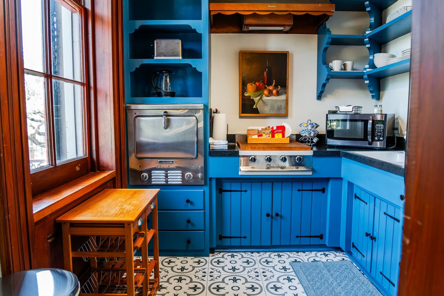 Cozy kitchen interior with bright blue cabinets, patterned tile floor, and wooden window trim and counter.