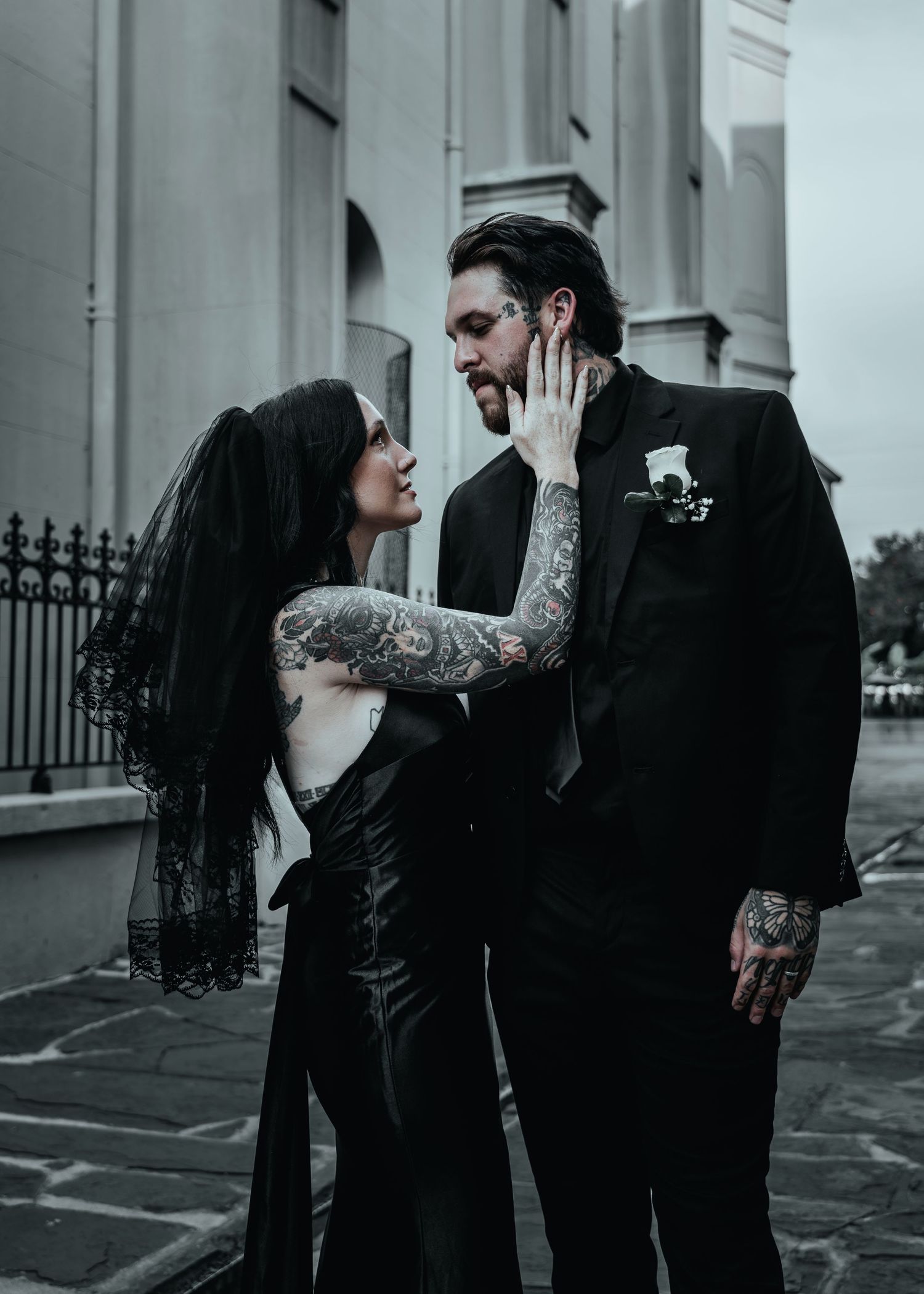 Gothic couple in black attire share an intimate moment outside historic columns in a moody monochromatic scene.
