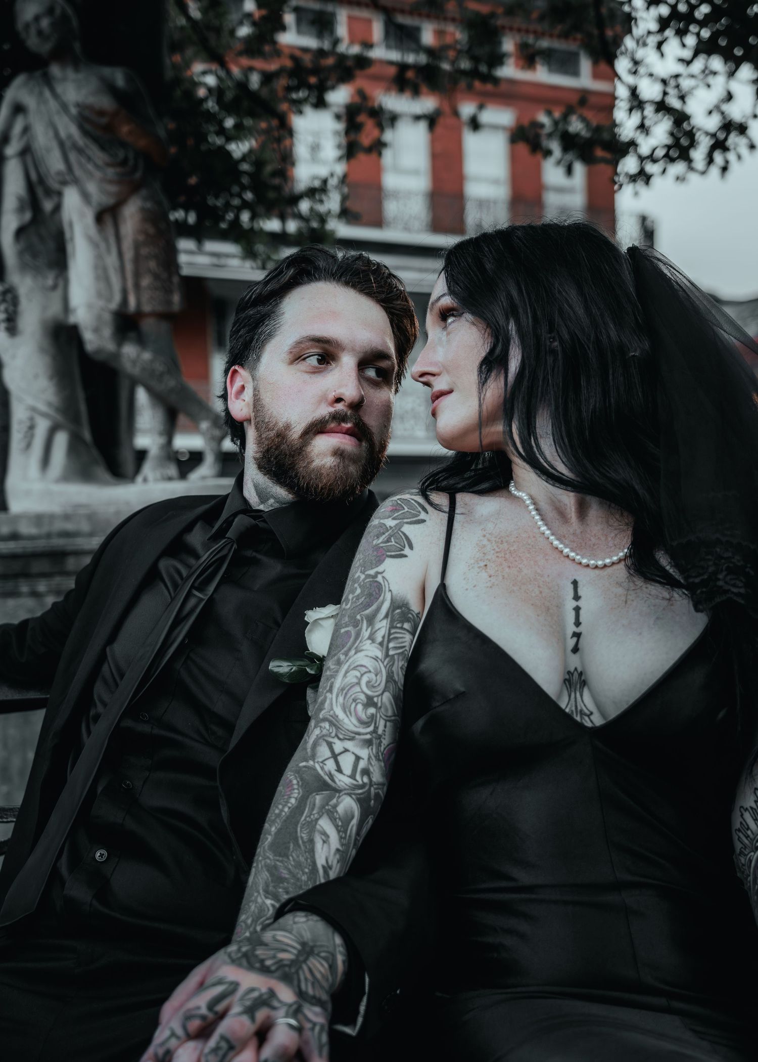 Gothic styled couple in black clothing embracing against street backdrop with vintage buildings visible.