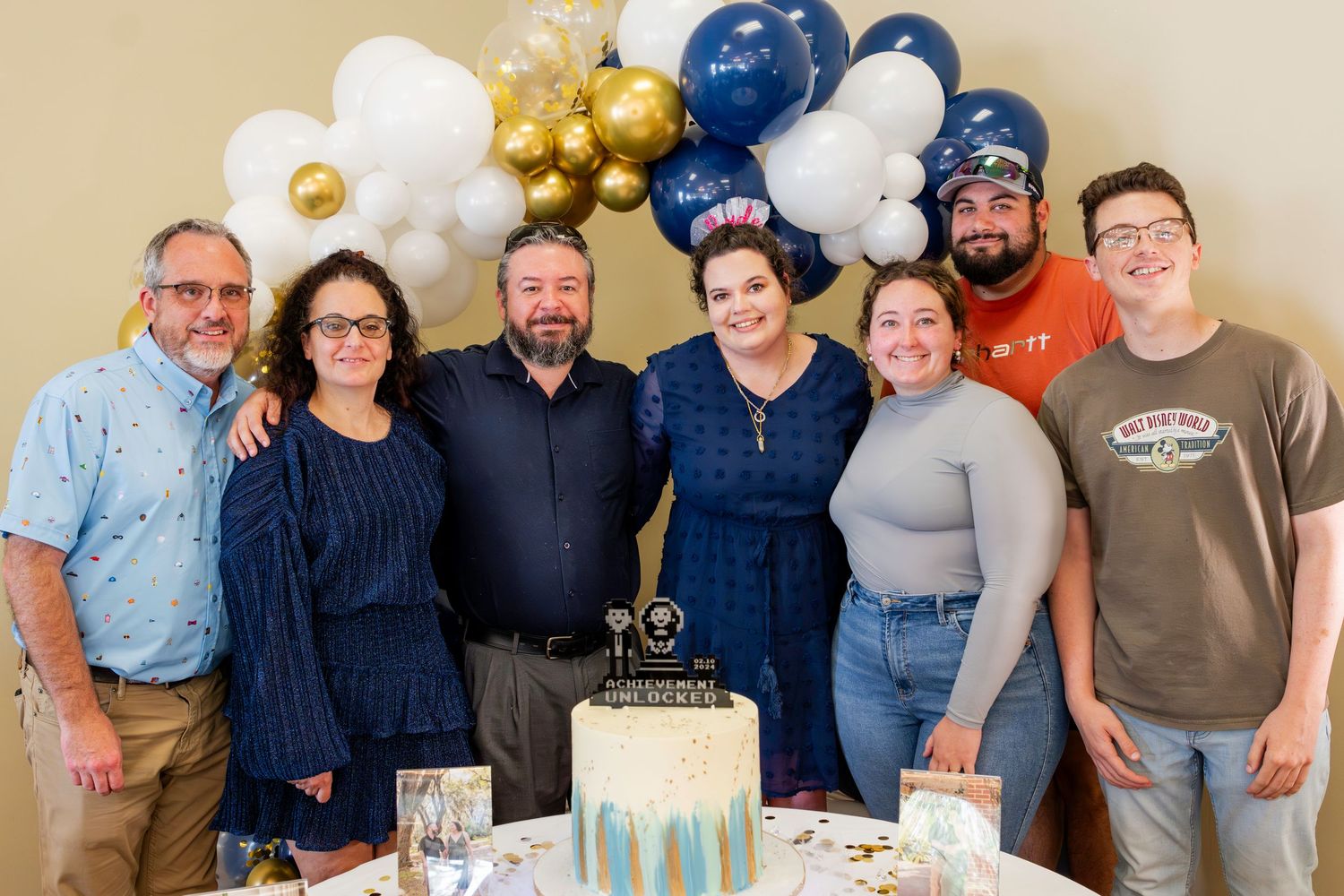 Group celebrates special occasion with gold and blue balloon decorations and a cake at casual indoor gathering.