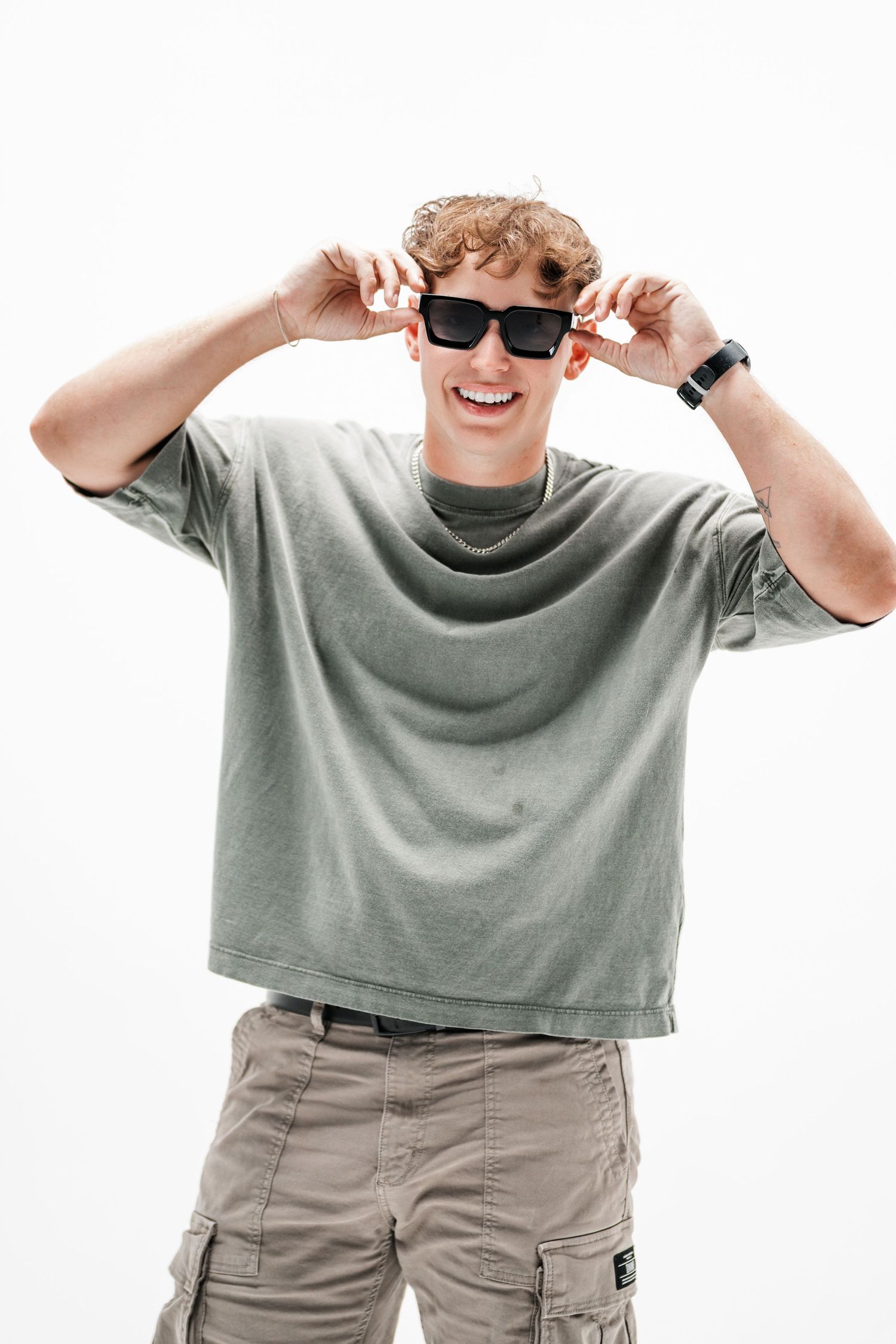 Young person in a gray t-shirt and cargo pants adjusting sunglasses while smiling.