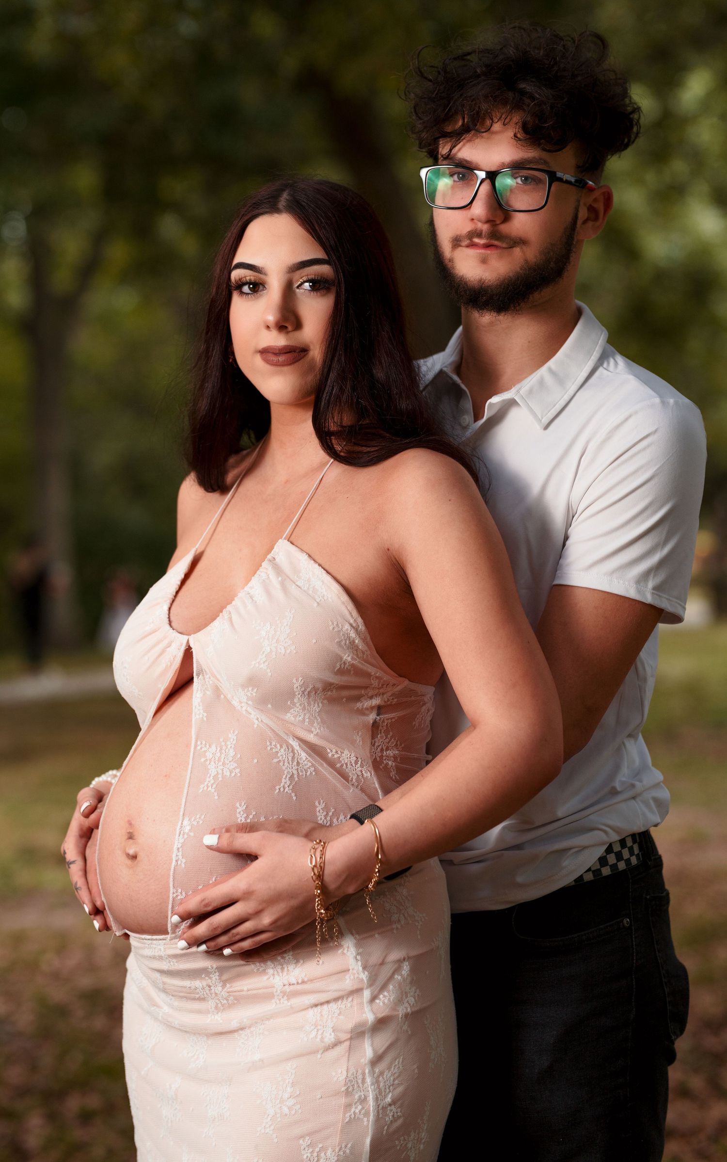 A couple poses outdoors during a maternity photoshoot, with the expectant mother wearing a white dress against a natural backdrop.