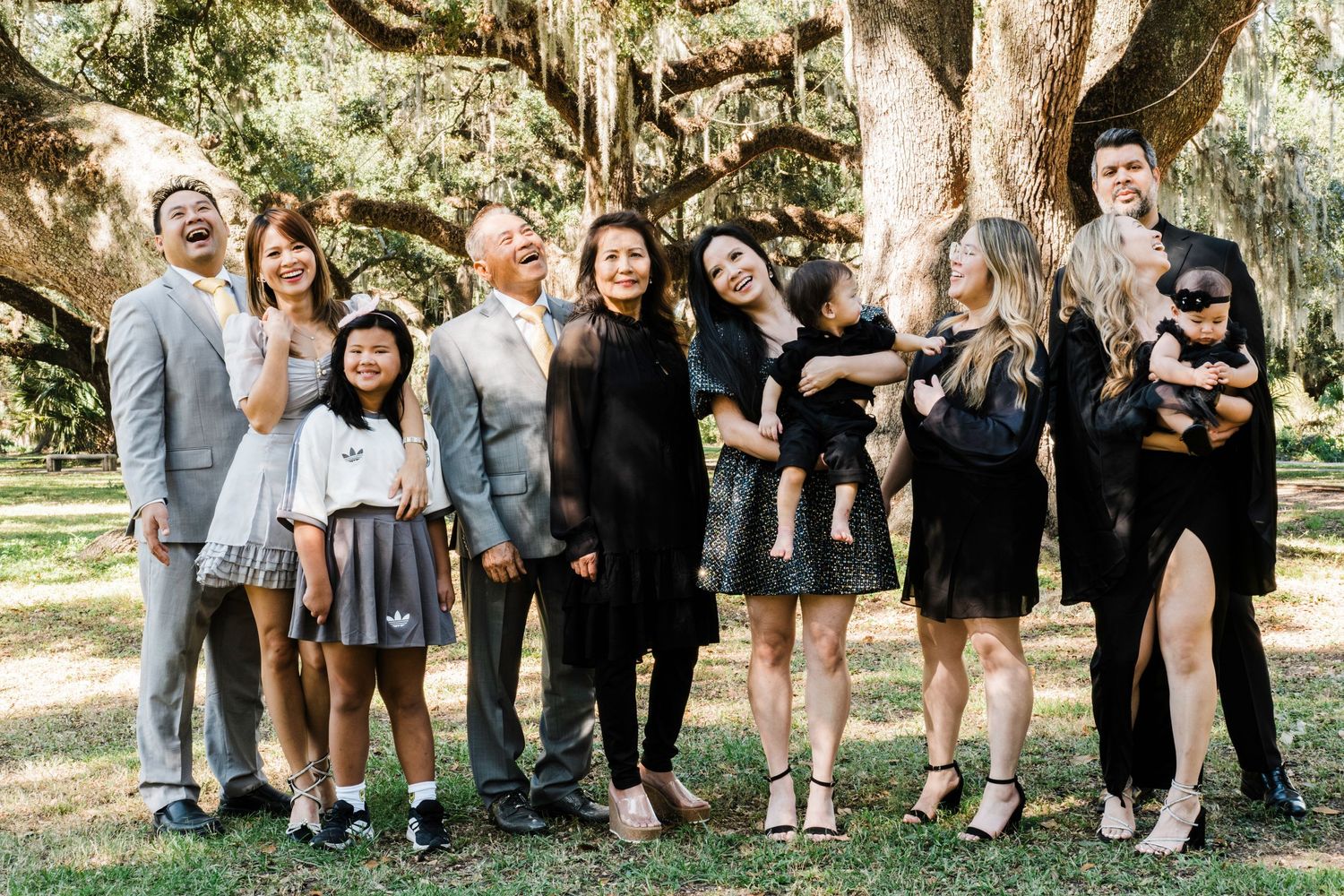 A large group of people dressed in formal attire pose together under moss-draped trees.