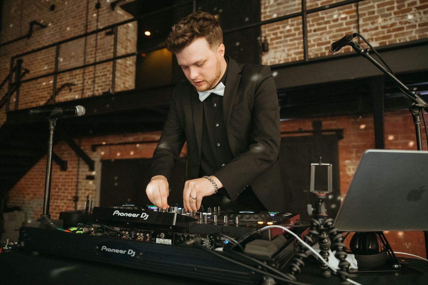 A DJ performs on Pioneer DJ equipment in front of a brick wall at an indoor venue.