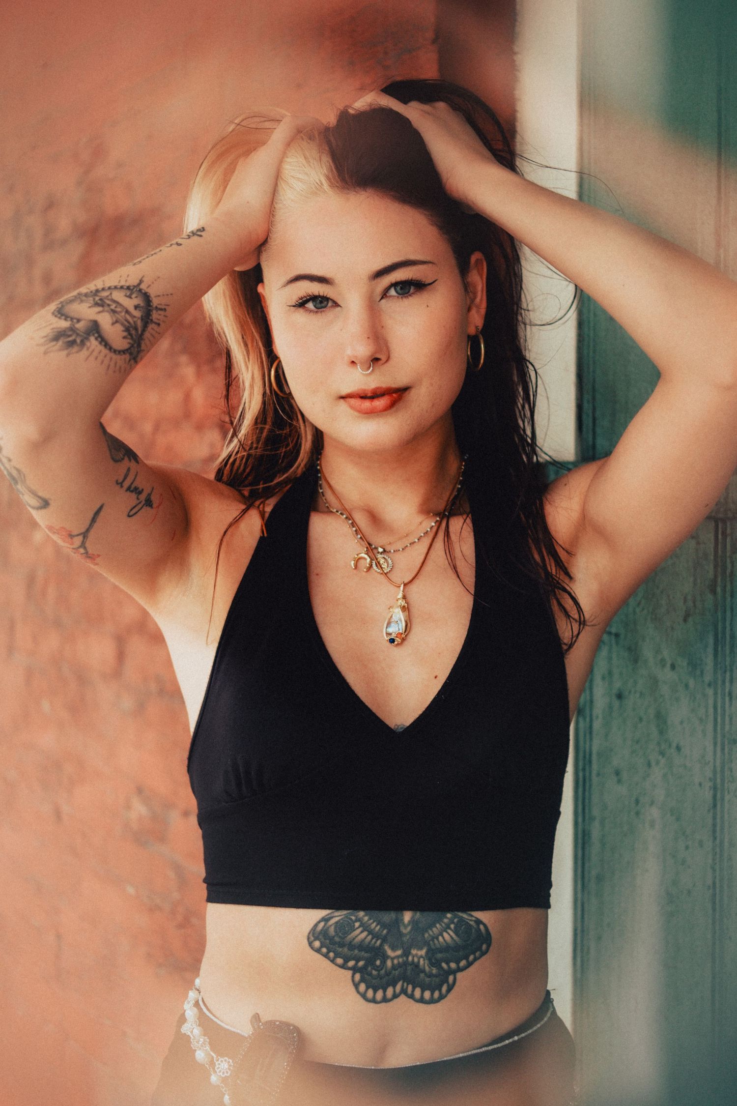 Portrait of model in black crop top with butterfly tattoo and pendant necklace against pink and green wall.