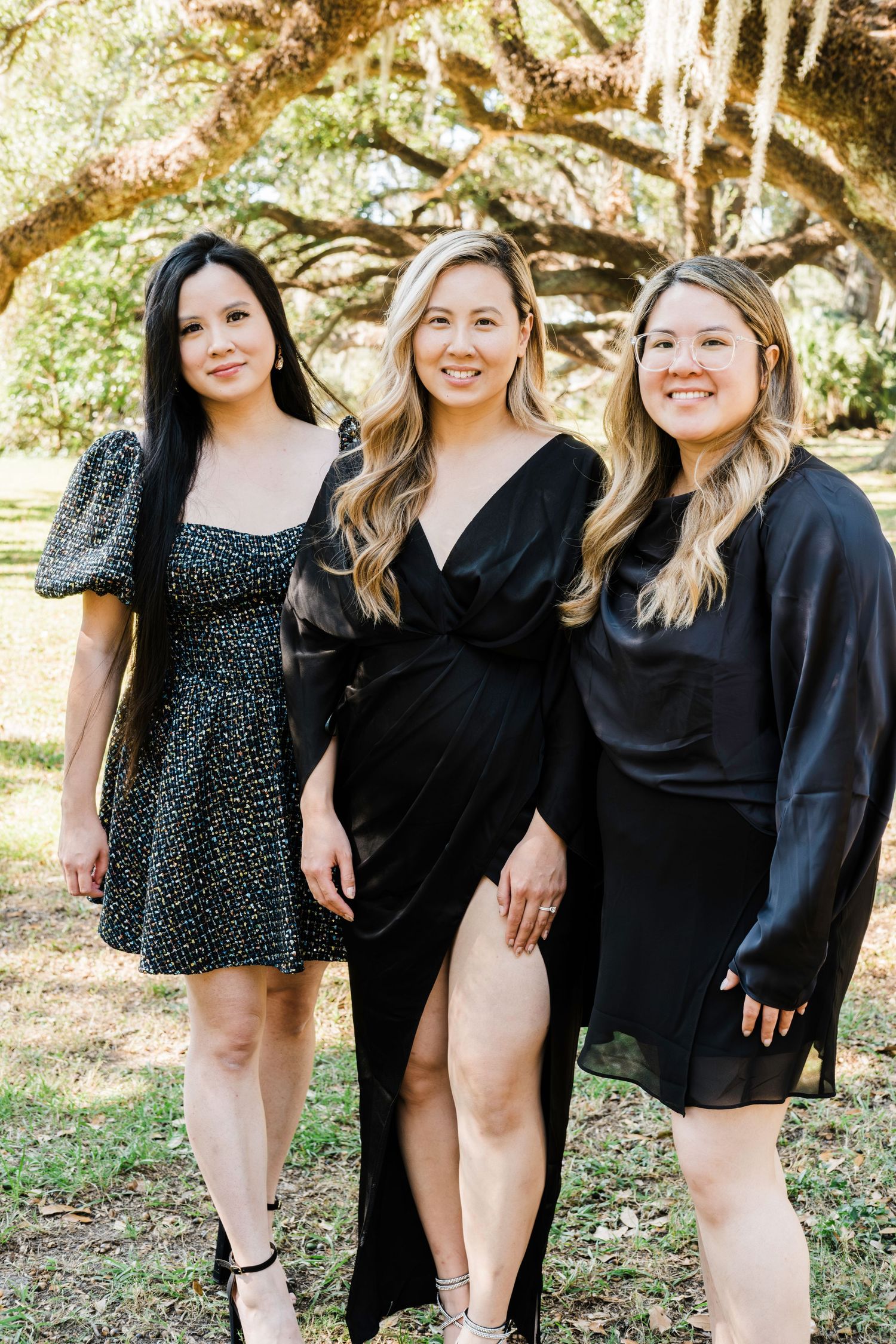 Three friends wearing black dresses stand together under oak trees in a sunlit outdoor setting.