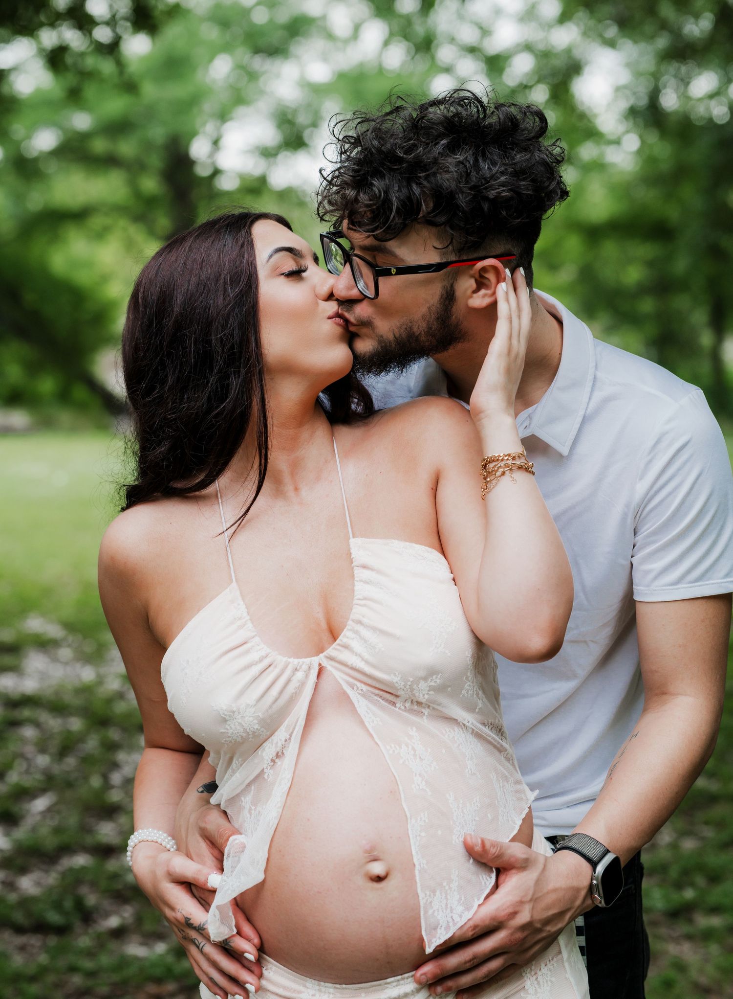 An expecting couple shares a tender kiss in a lush garden setting while gently cradling her pregnant belly.